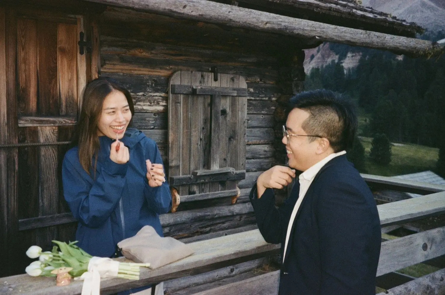 Couple laughing by a wooden mountain hut after a proposal, on 35mm film