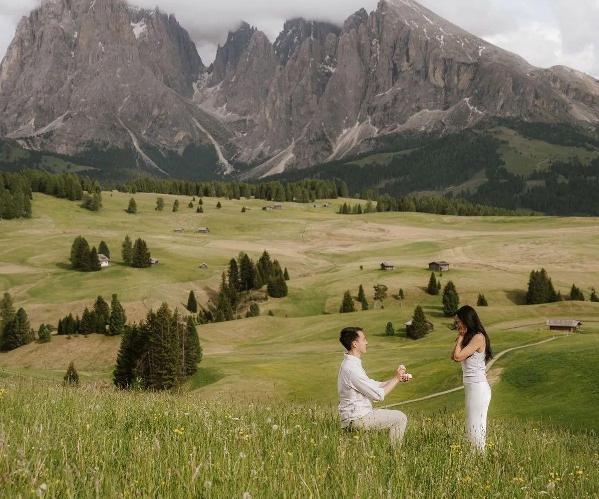 Man kneels to propose in a wide meadow on the Alpe di Siusi, Sassolungo peaks behind