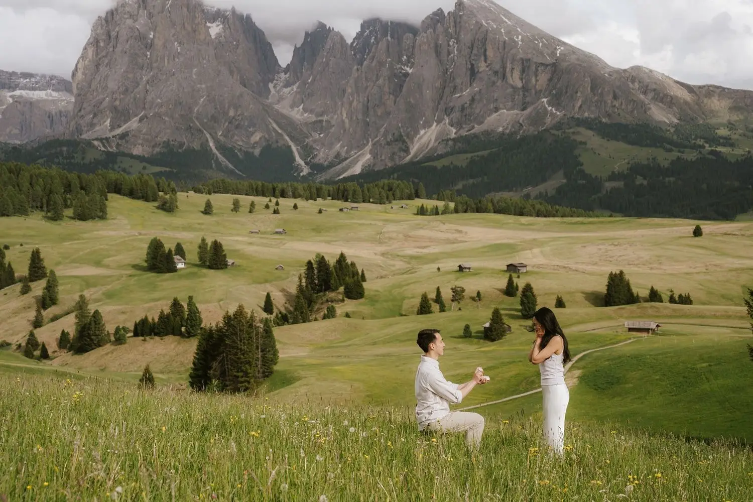 Man kneels to propose in a wide meadow on the Alpe di Siusi, Sassolungo peaks behind
