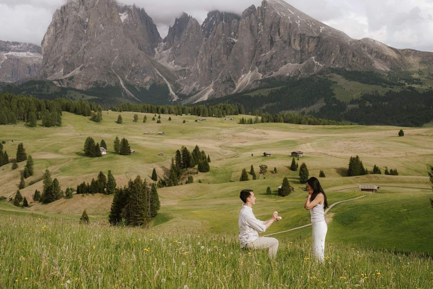 Man kneels to propose in a wide meadow on the Alpe di Siusi, Sassolungo peaks behind