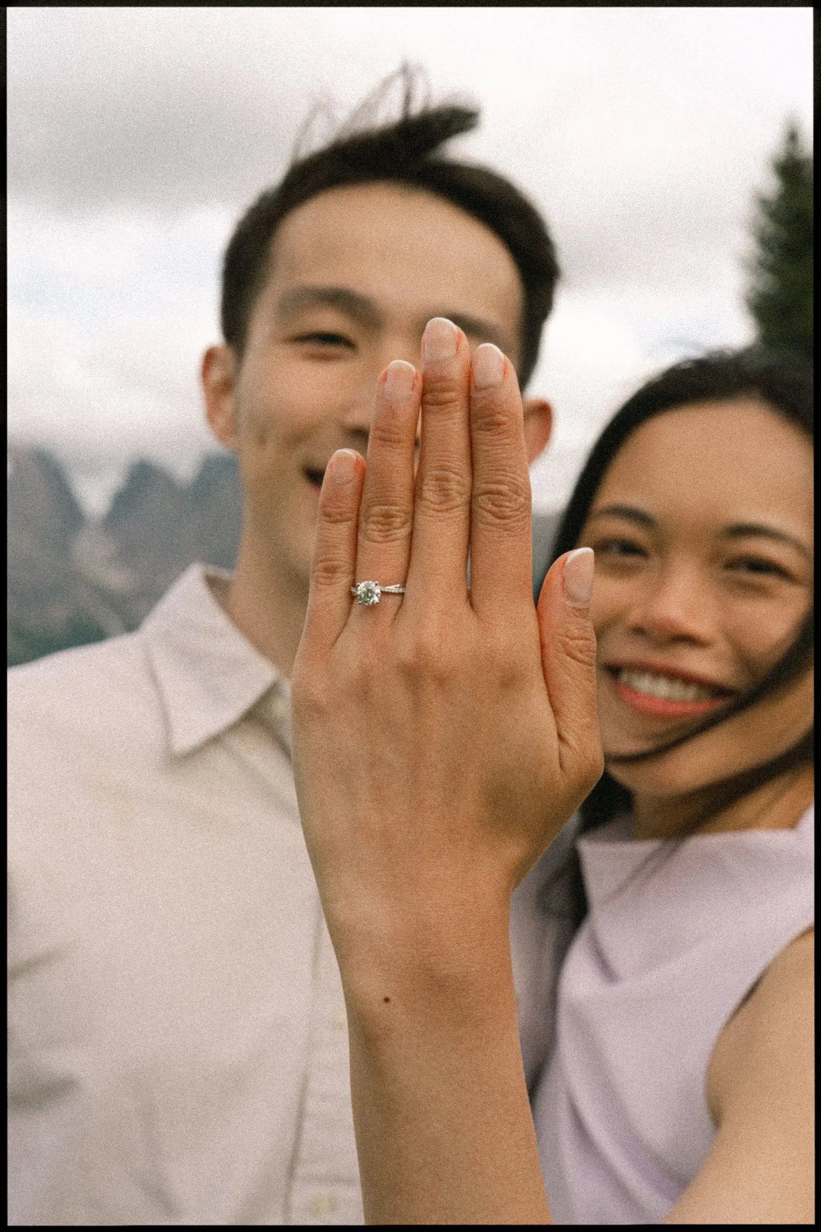Laughing fiancée shows her engagement ring to the camera, her partner beside her