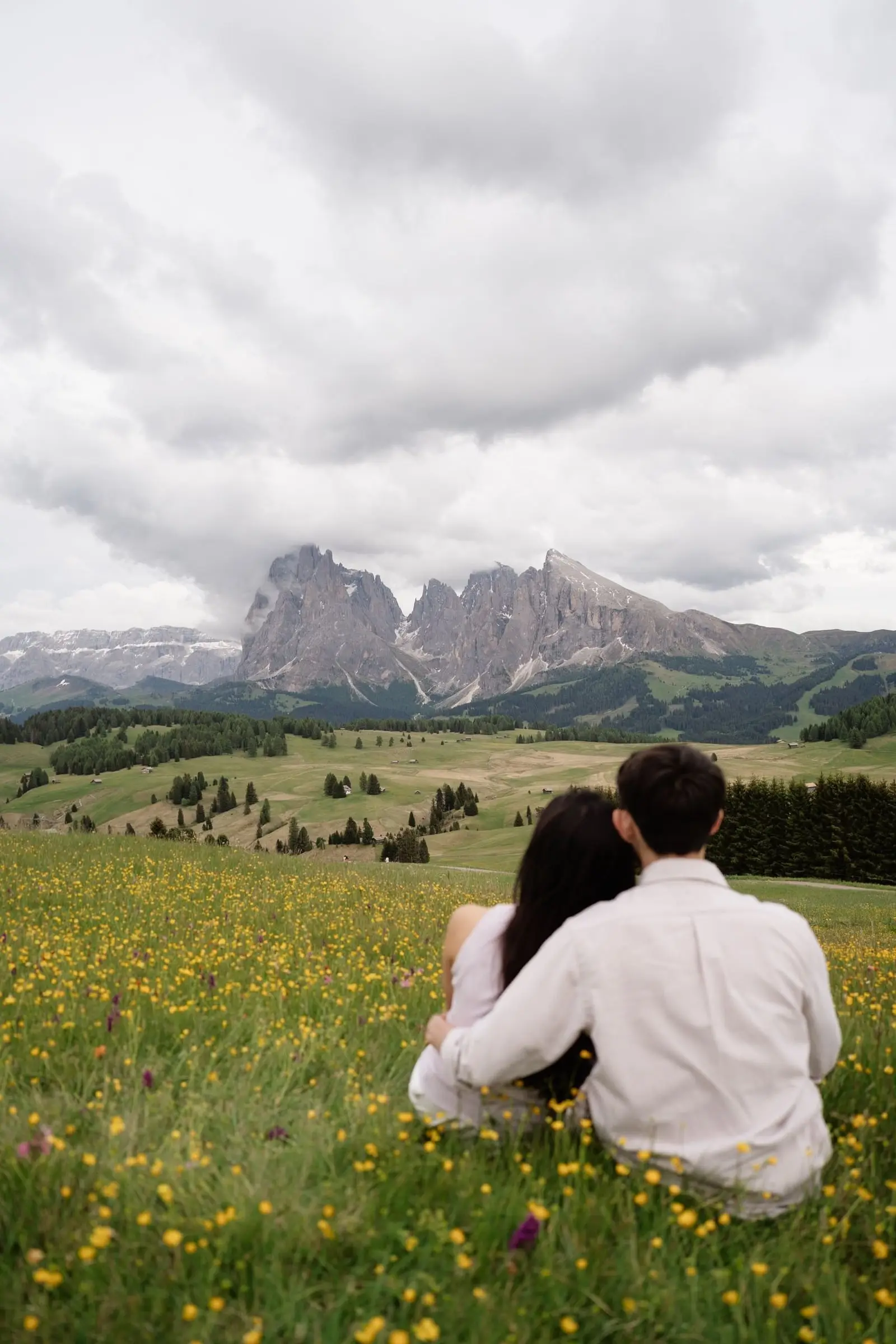 Couple sitting in a field of yellow flowers, looking towards the Sassolungo