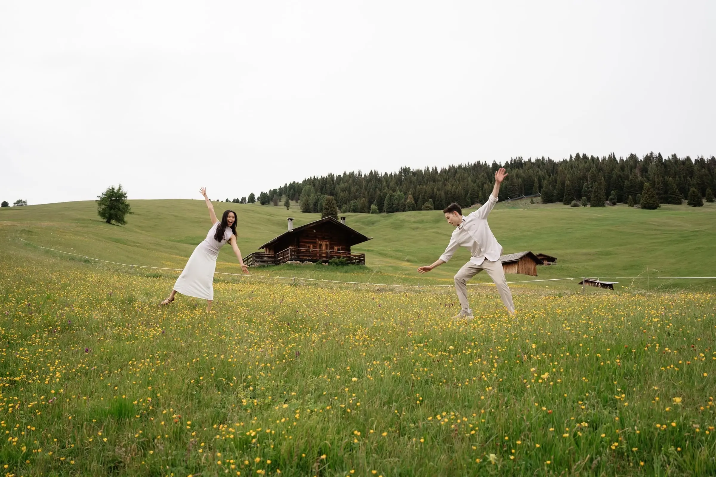 Couple jumping playfully in a flower meadow next to a wooden hut