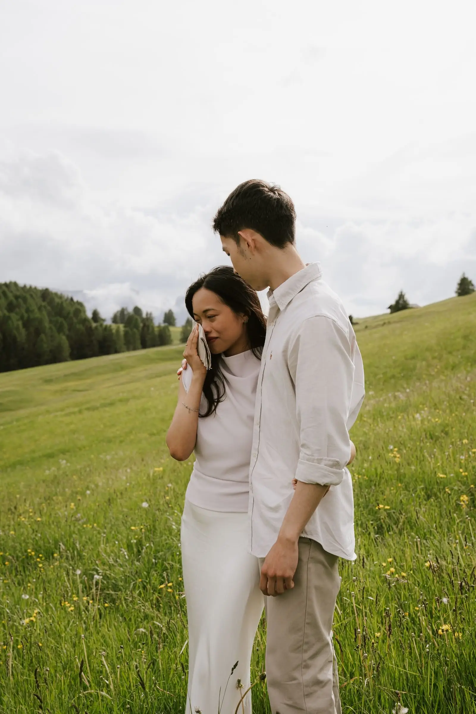 He kisses her forehead as she wipes a tear in the meadow