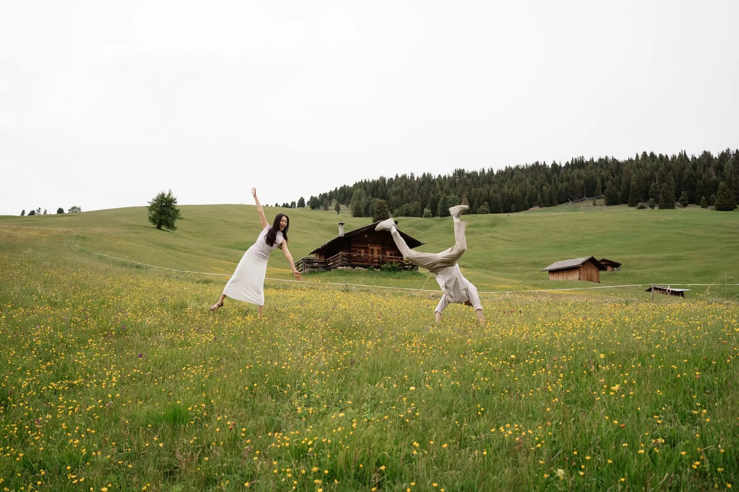 He does a cartwheel while she laughs in the meadow next to a hut