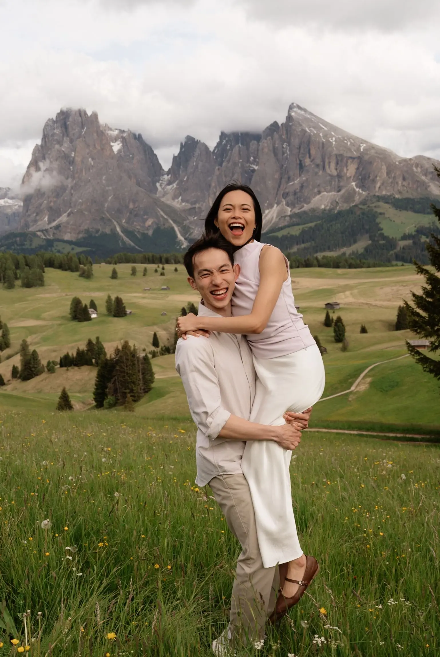He carries her on his back, both laughing, below the Sassolungo on the Alpe di Siusi