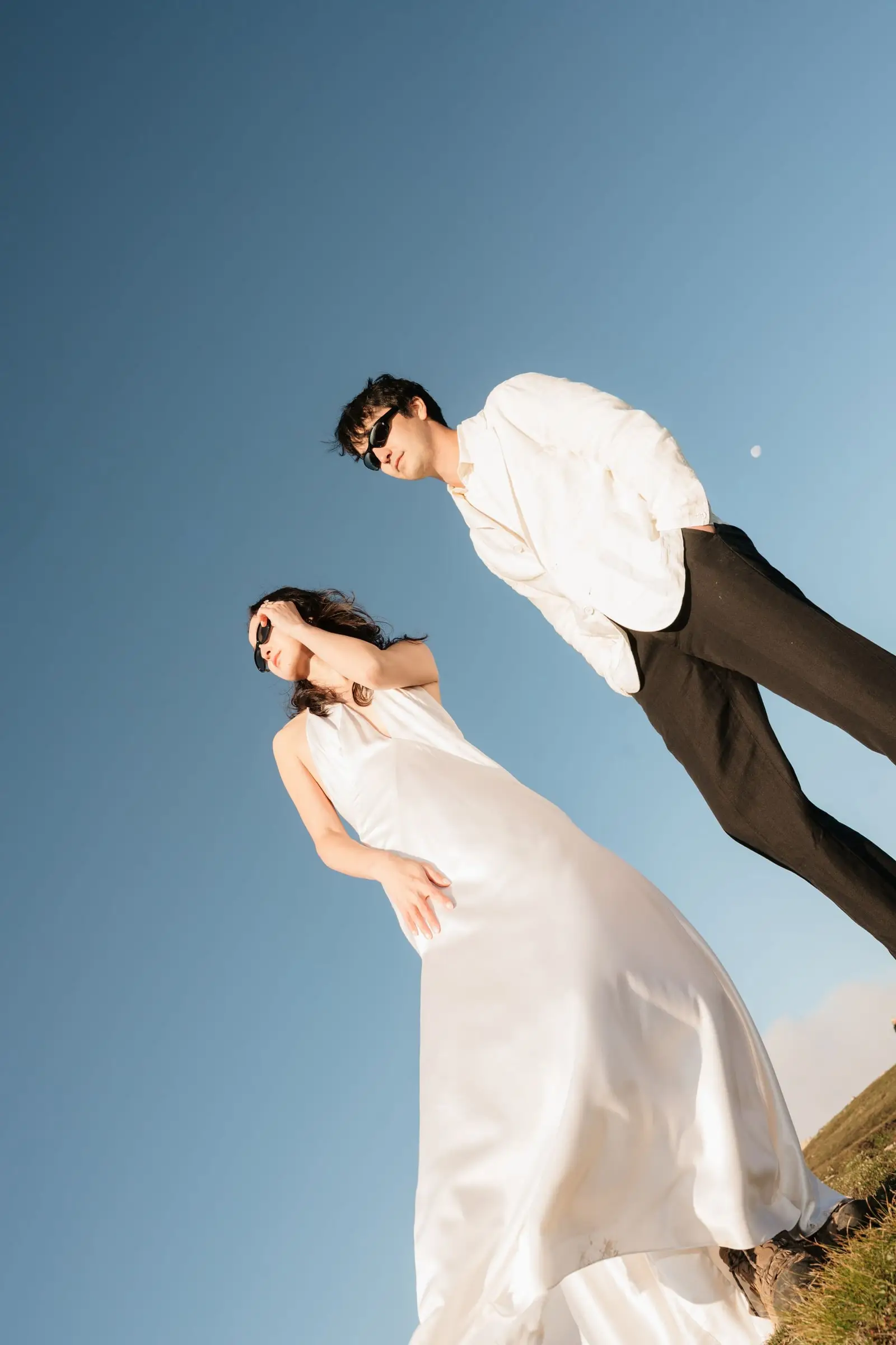 Couple in a silk gown and white jacket leaning into the frame against a clear blue sky