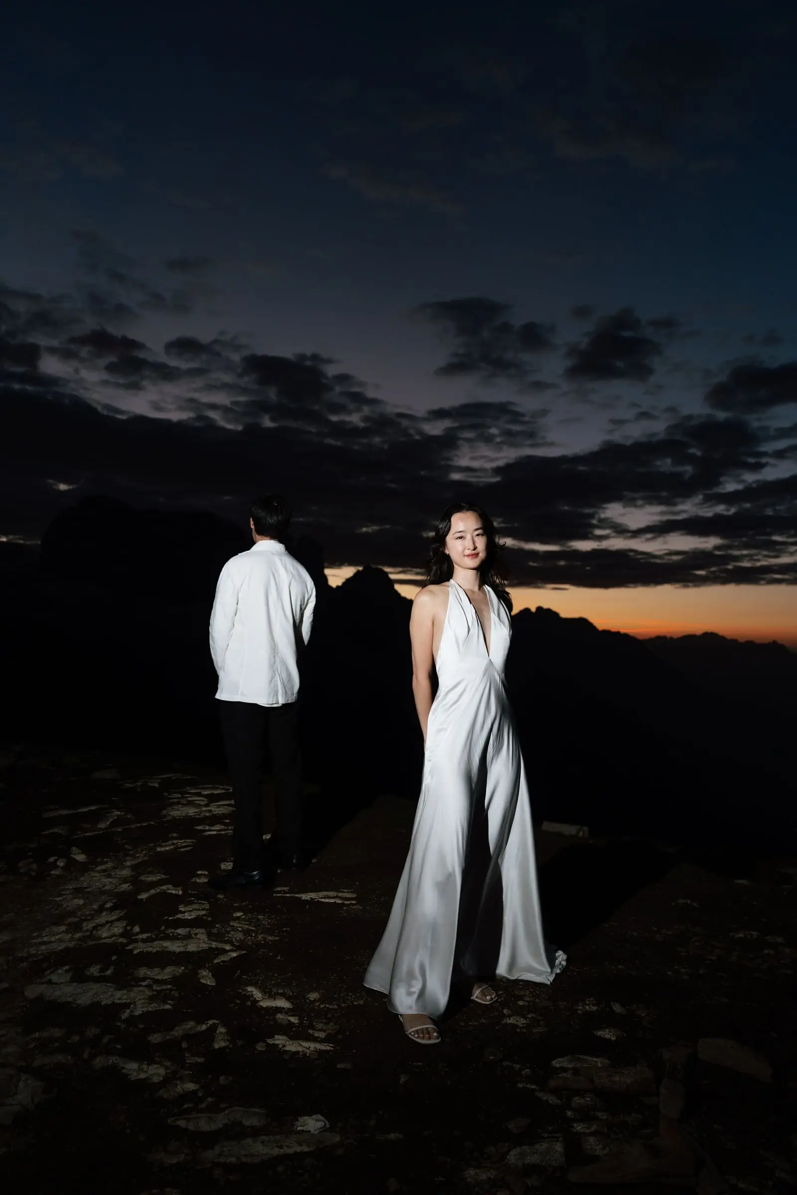 Bride in a silk gown and groom on a dark mountain ridge before dawn