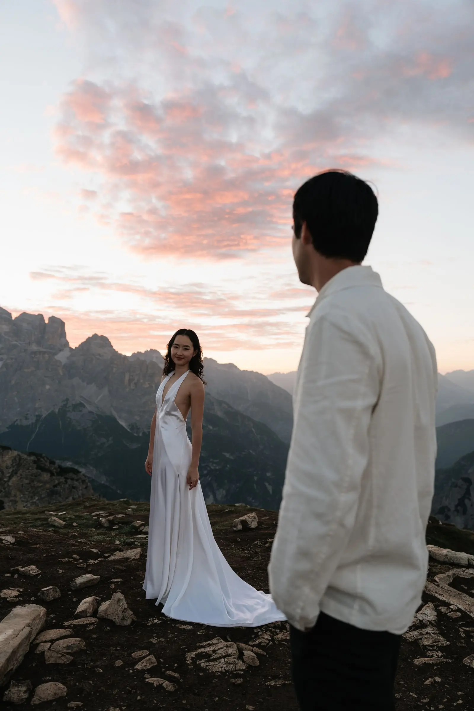 Bride looks back at the groom under pink clouds above the Dolomites
