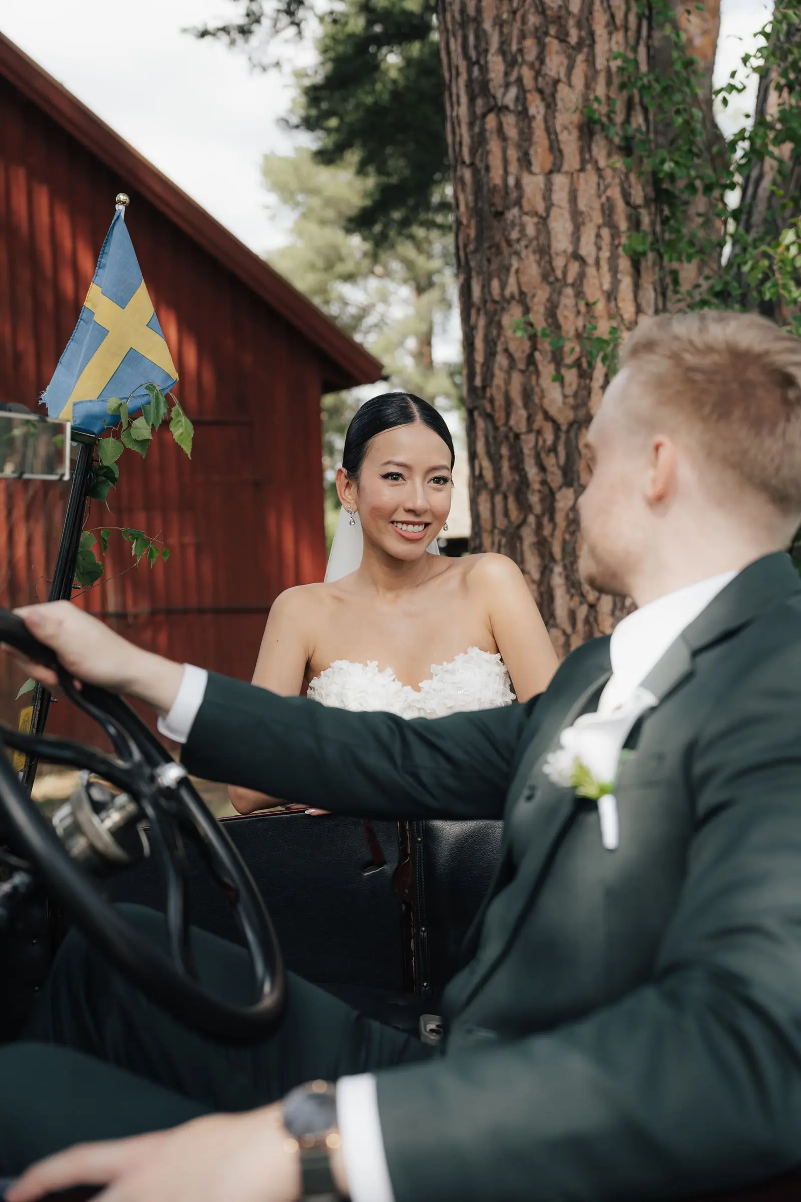 Bride smiles at the groom at the wheel of a vintage car beside a red Swedish house