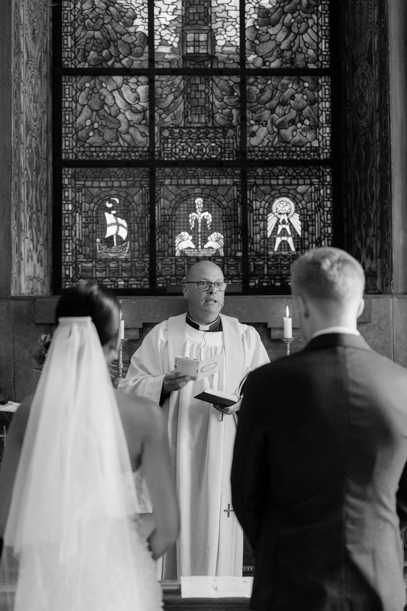 Black and white photo of the priest leading the ceremony before a stained-glass window