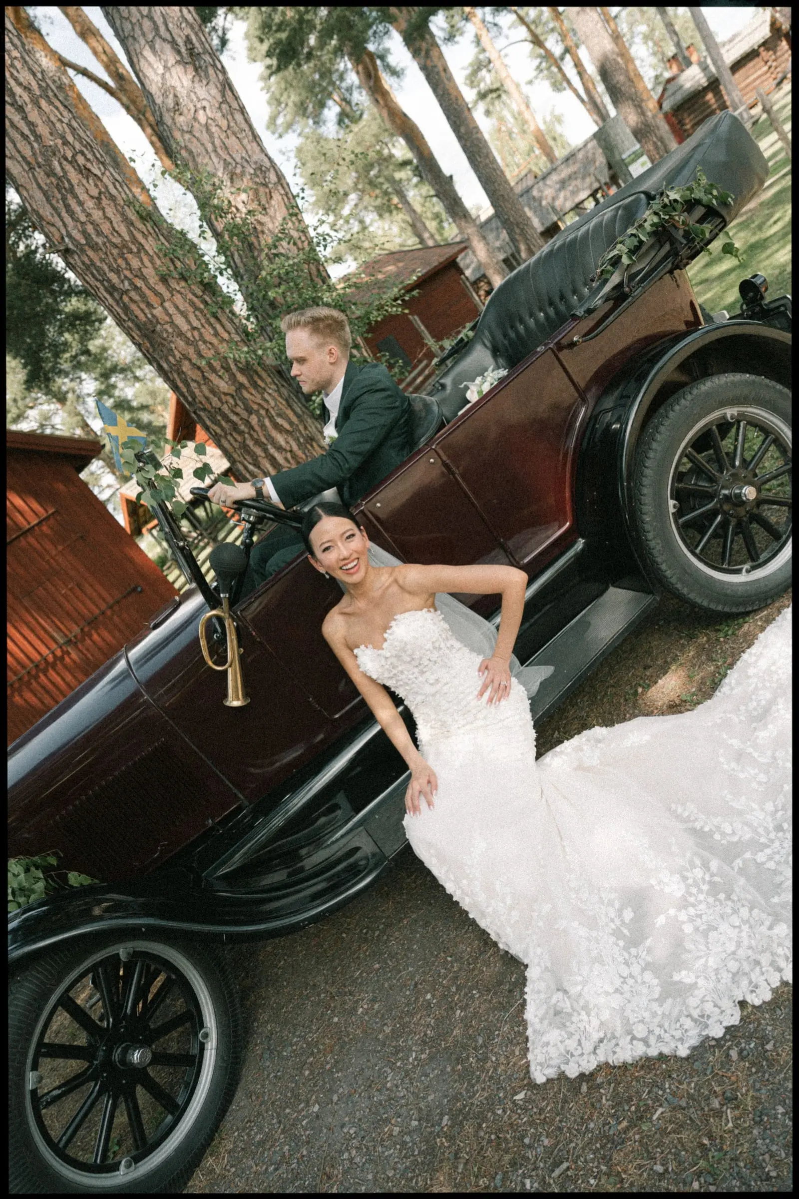 Bride posing beside a vintage car under tall pines, on film