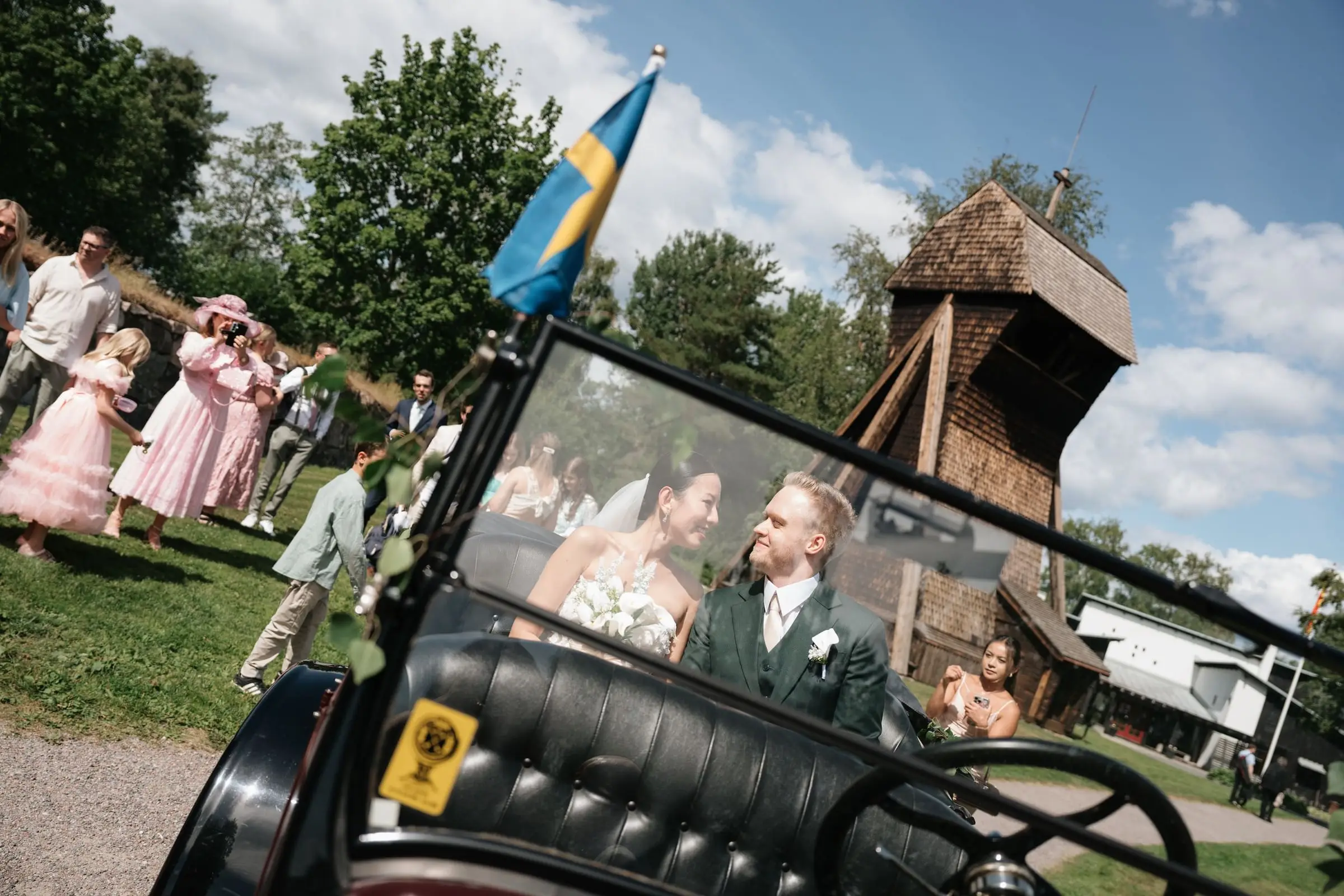 Newlyweds in a vintage car with a Swedish flag, guests behind