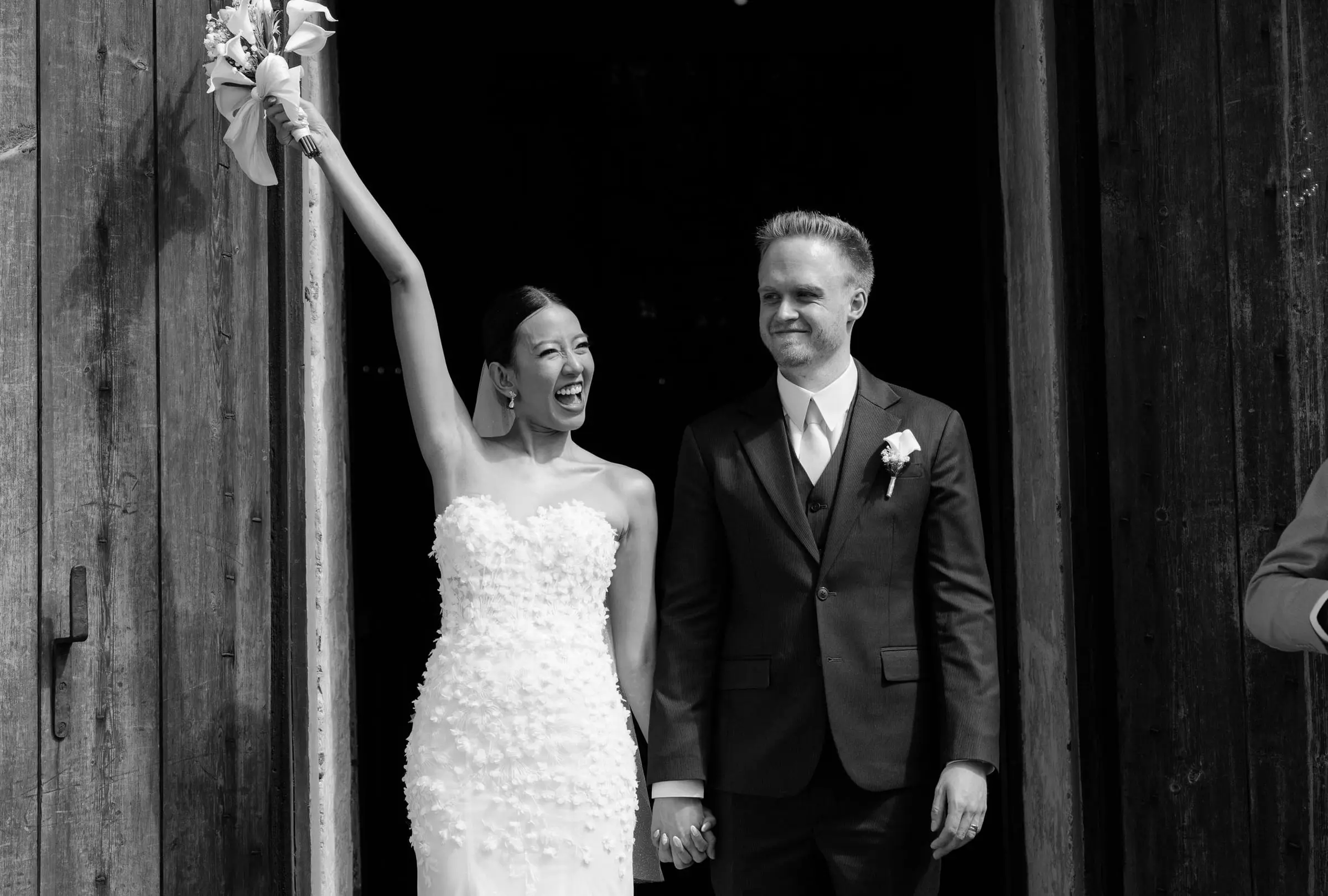 Black and white photo of the bride raising her bouquet as they leave the church