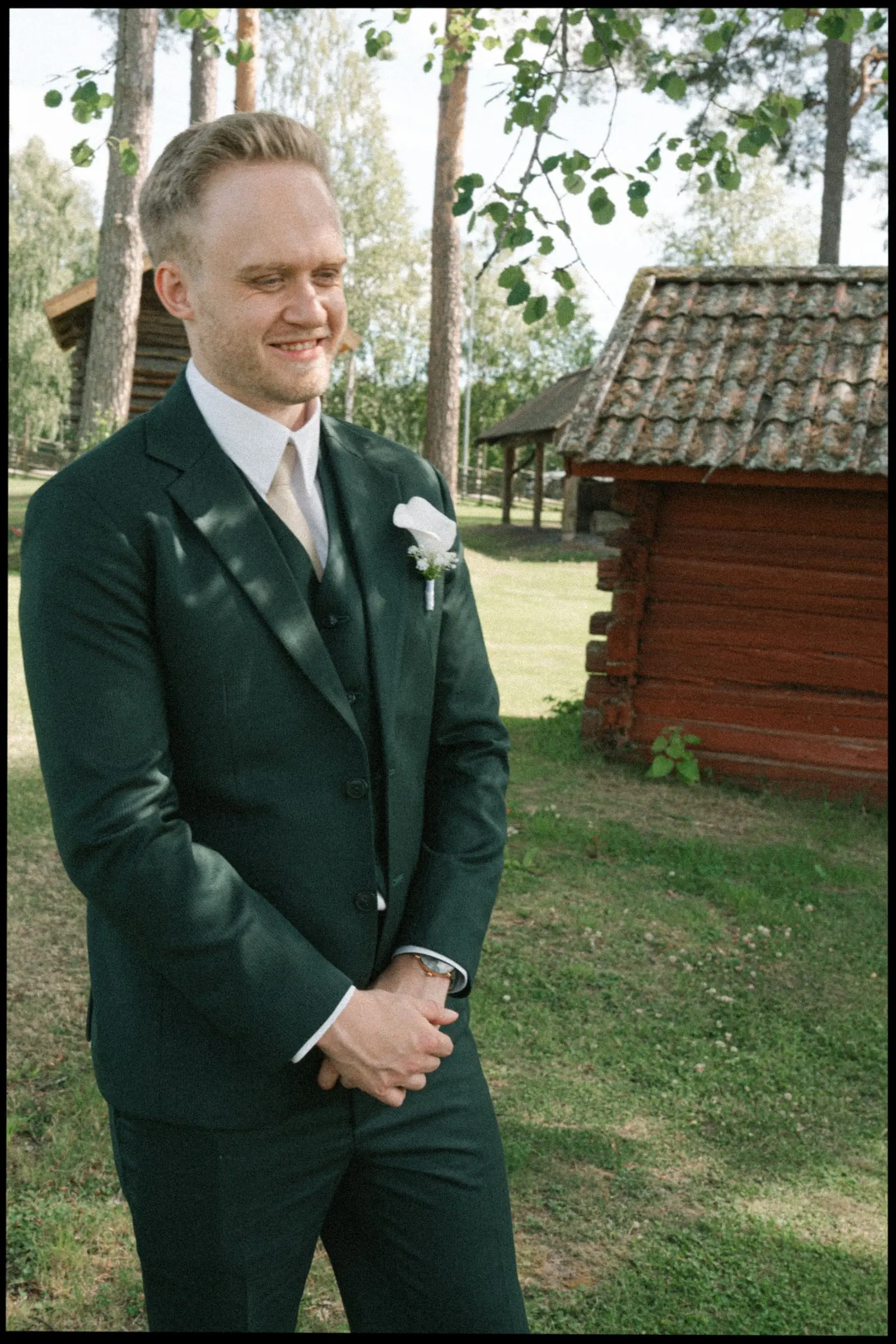 Smiling groom in a green suit by red wooden cottages, on film