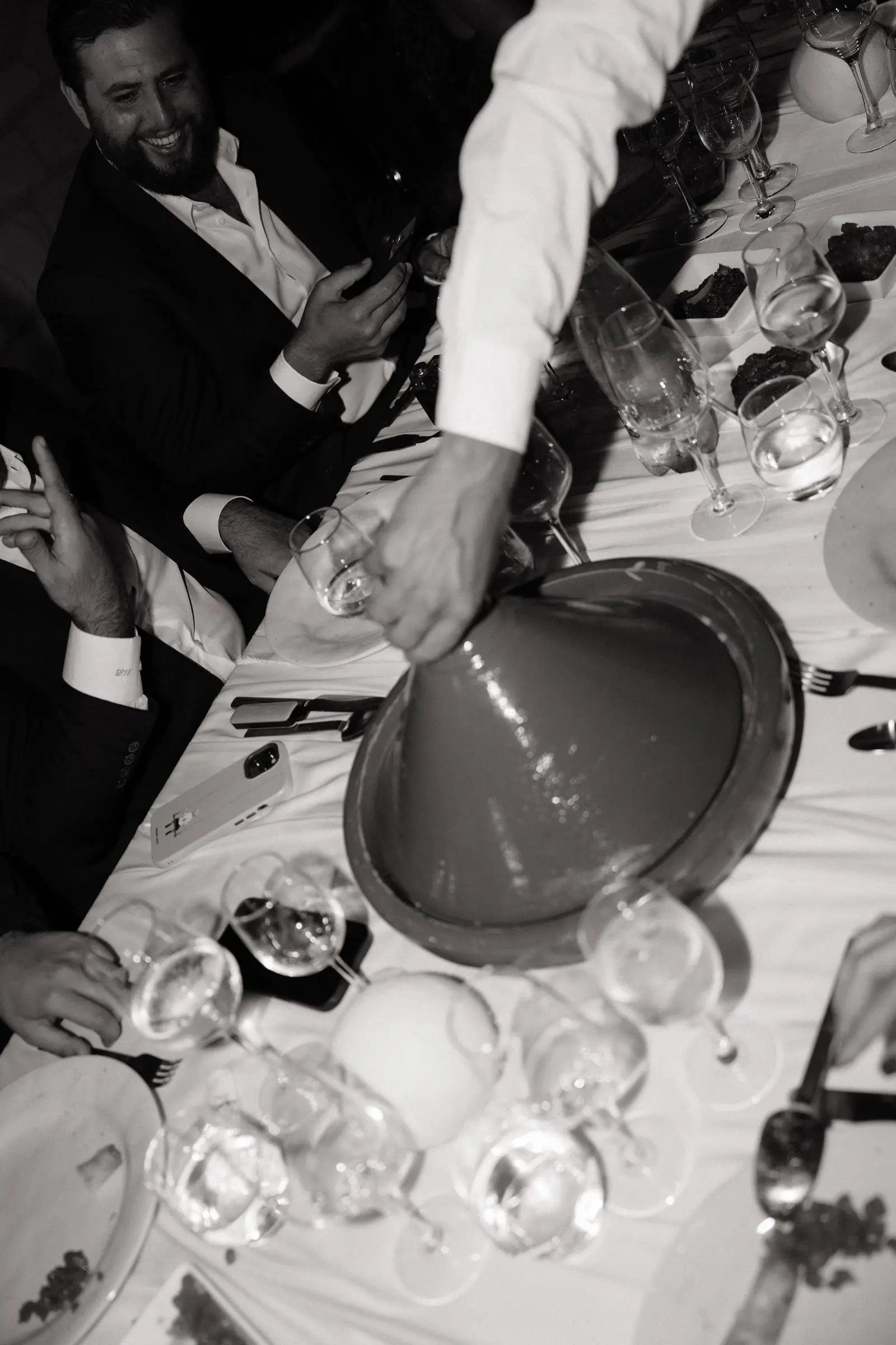 Black and white photo of a waiter lifting a tagine lid at the table