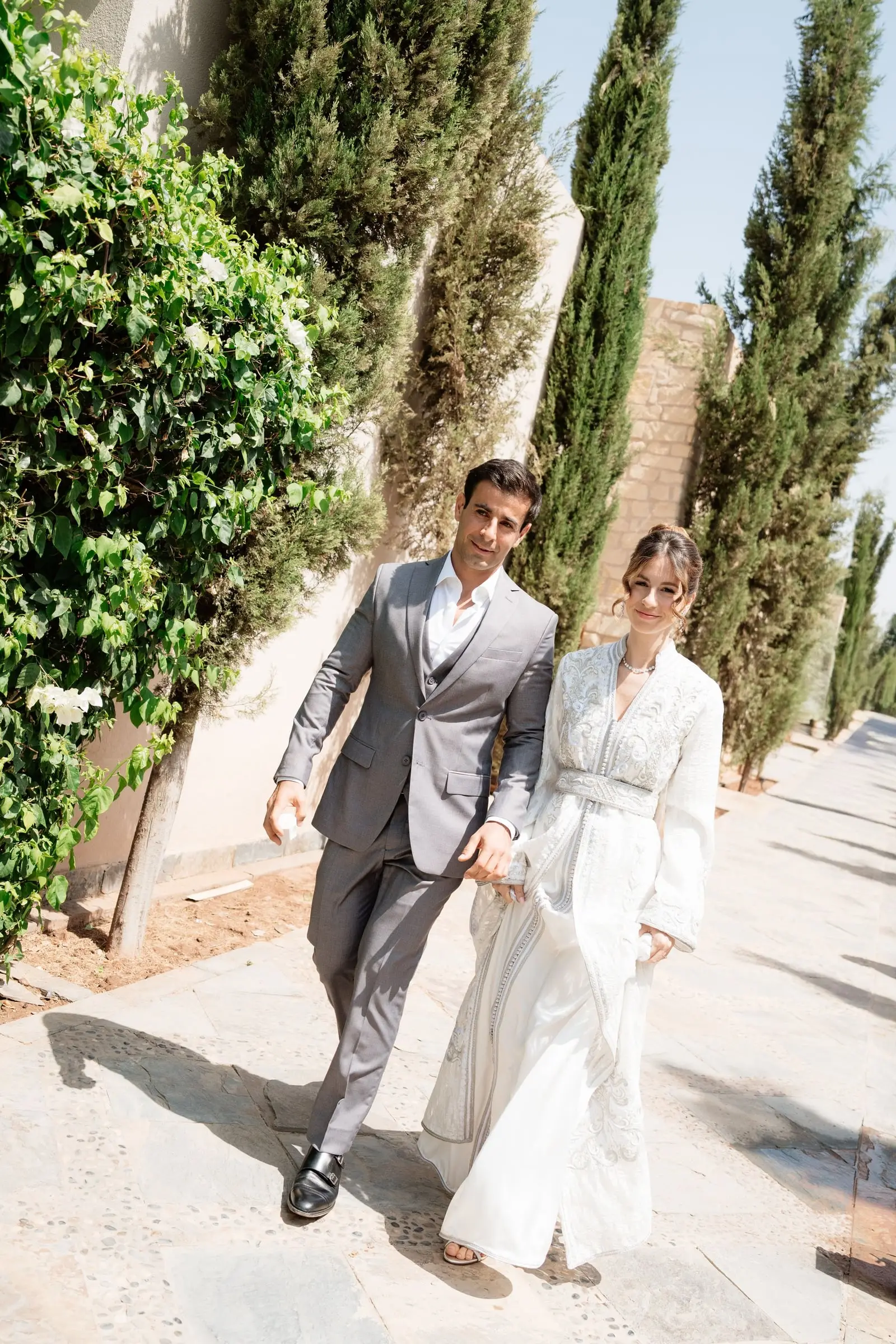Couple walking hand in hand past cypresses, the bride in a white embroidered kaftan