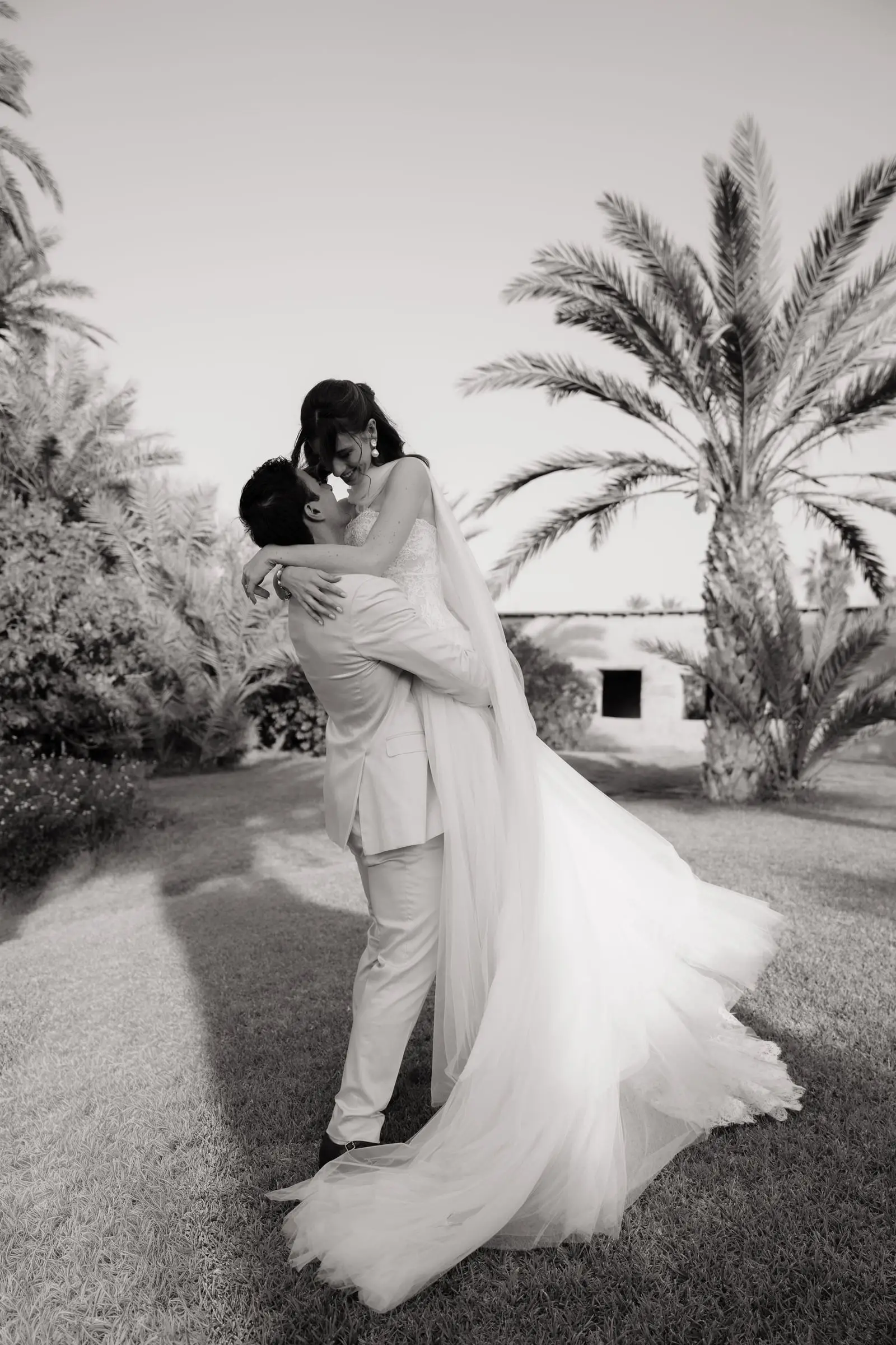 Black and white photo of the groom lifting the bride among palm trees