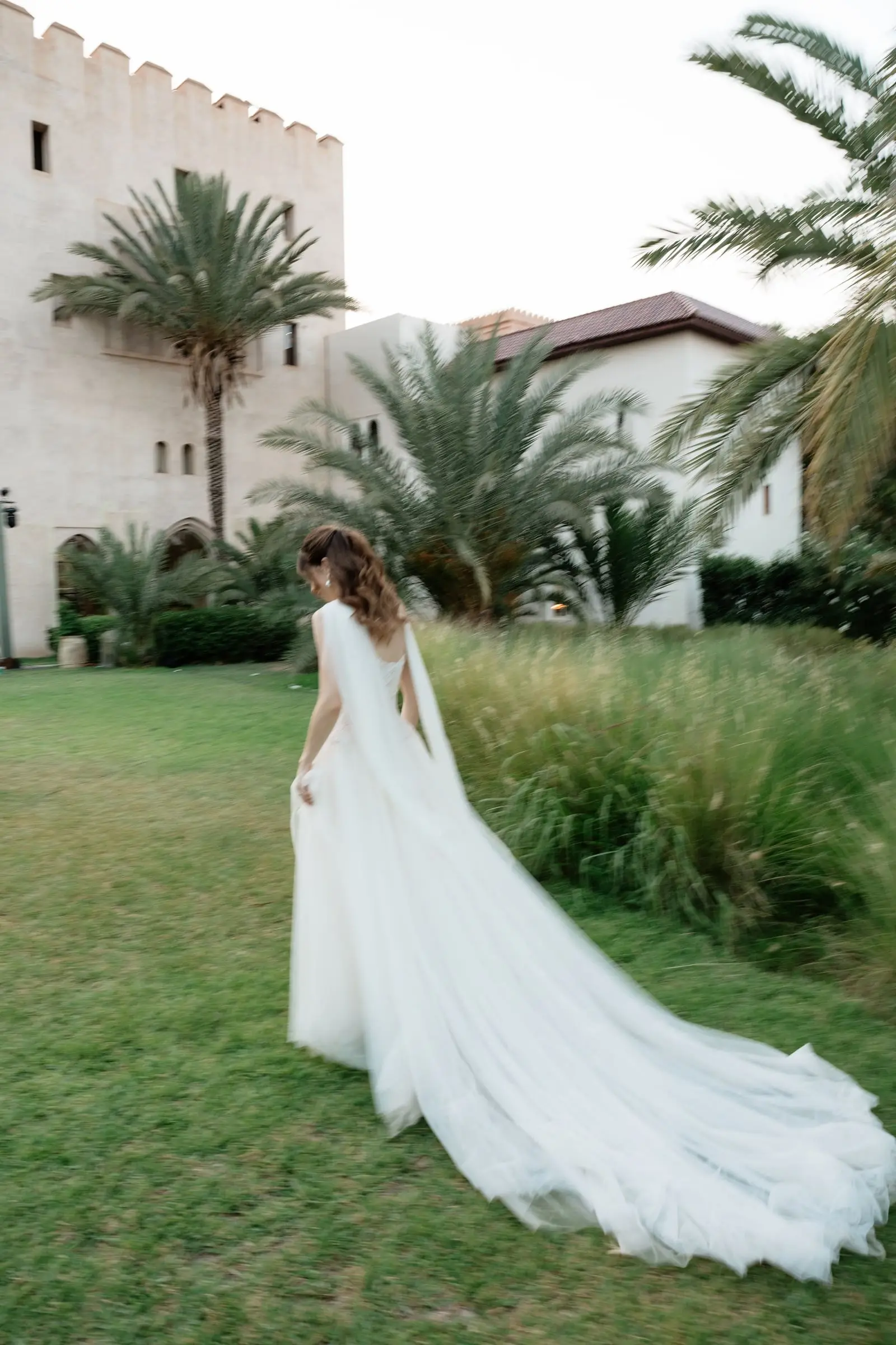 Bride with a long cape walking across the lawn towards the palace