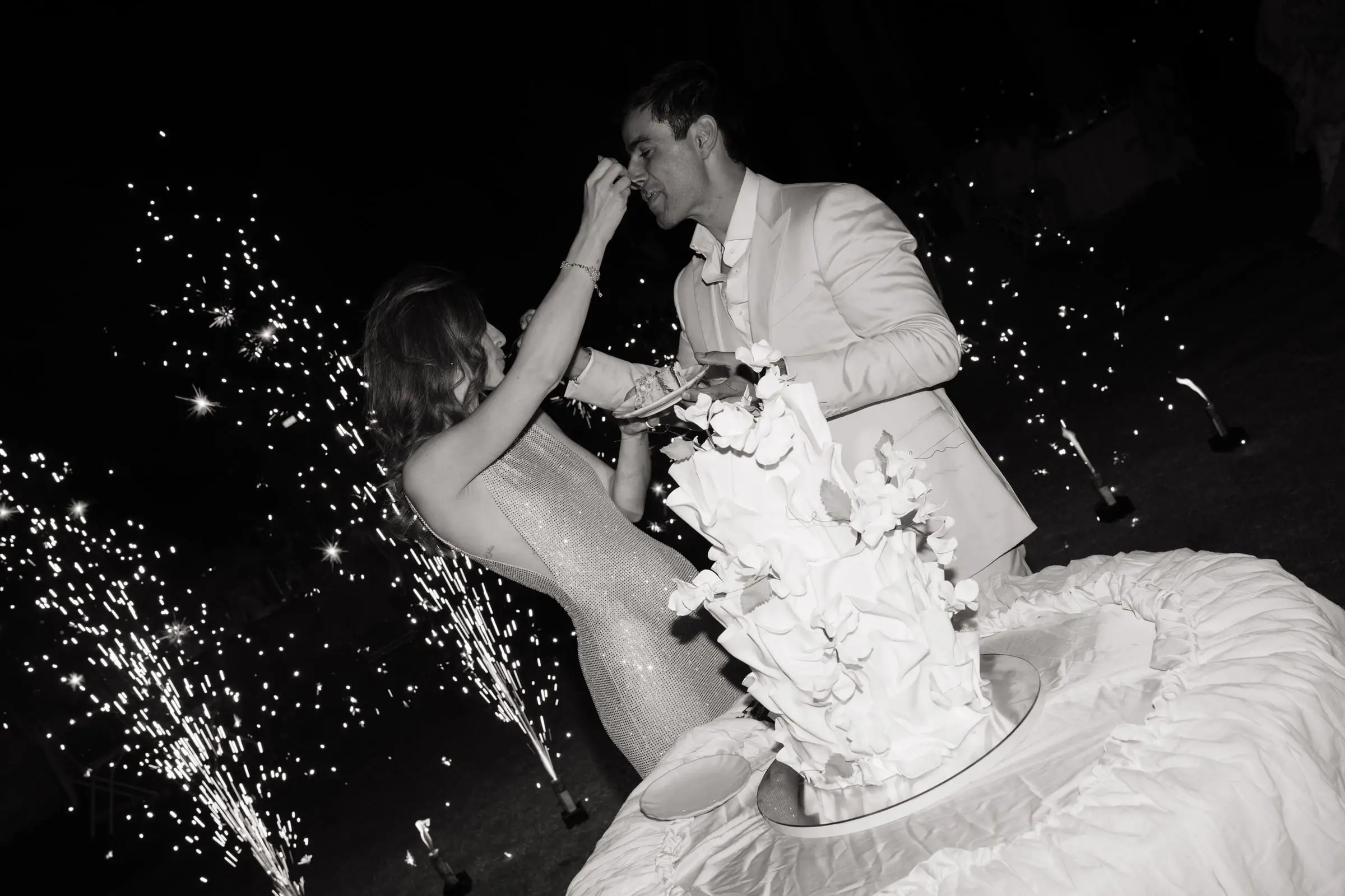 Black and white photo of the couple feeding each other cake among sparklers