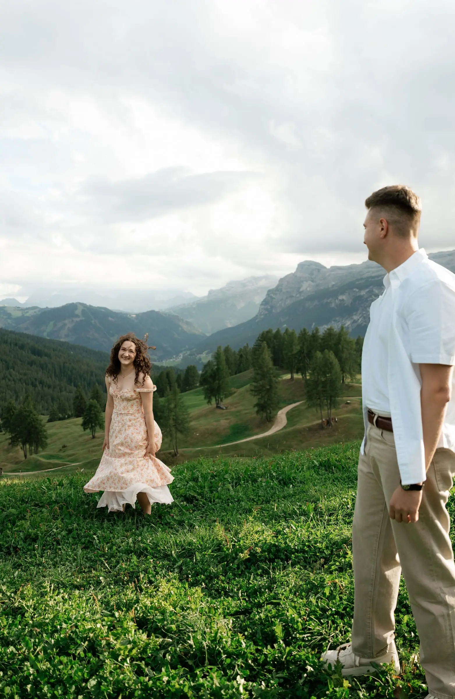 She walks towards him across an alpine meadow with mountains behind