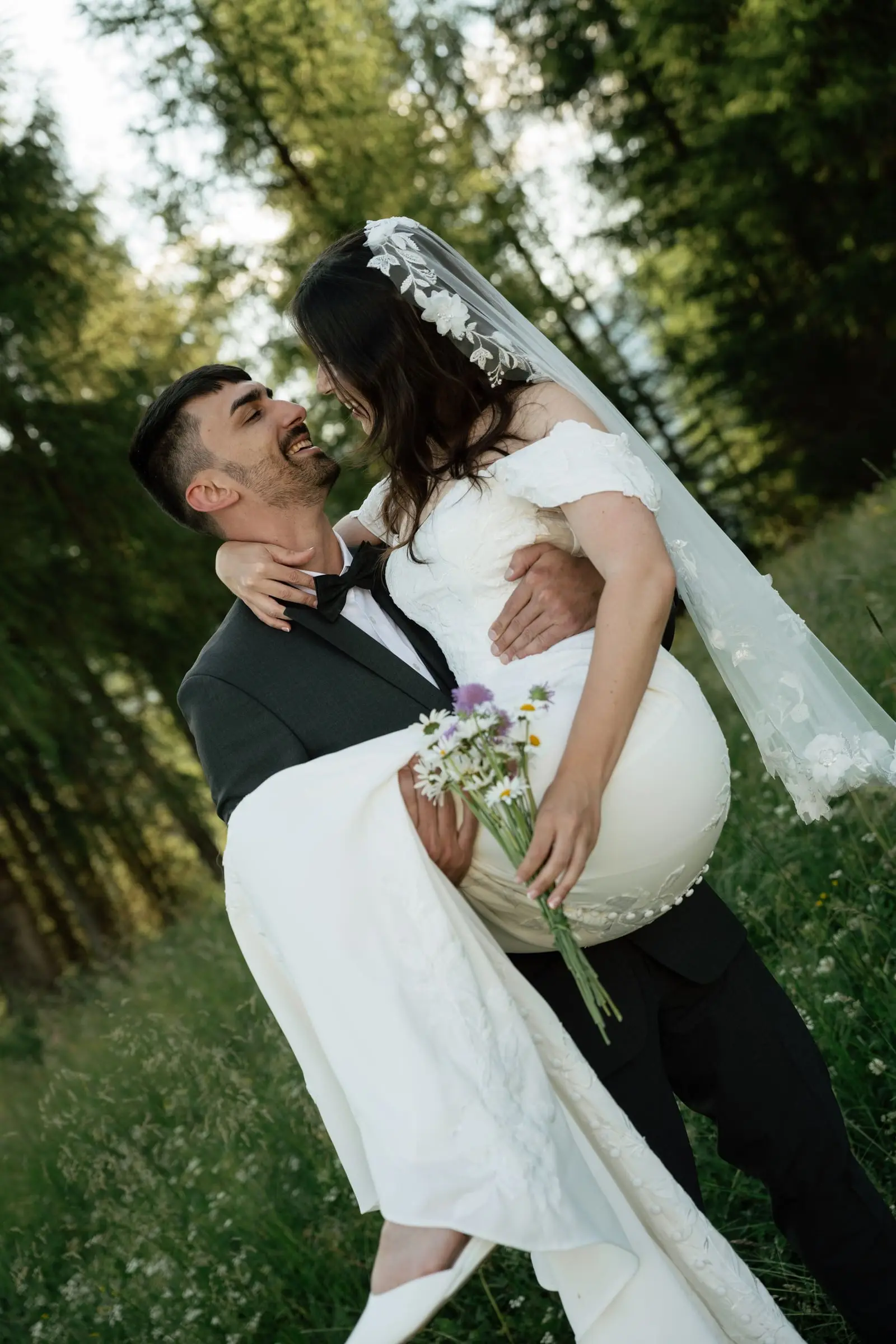 Groom lifts the laughing bride with her wildflower bouquet at the forest edge