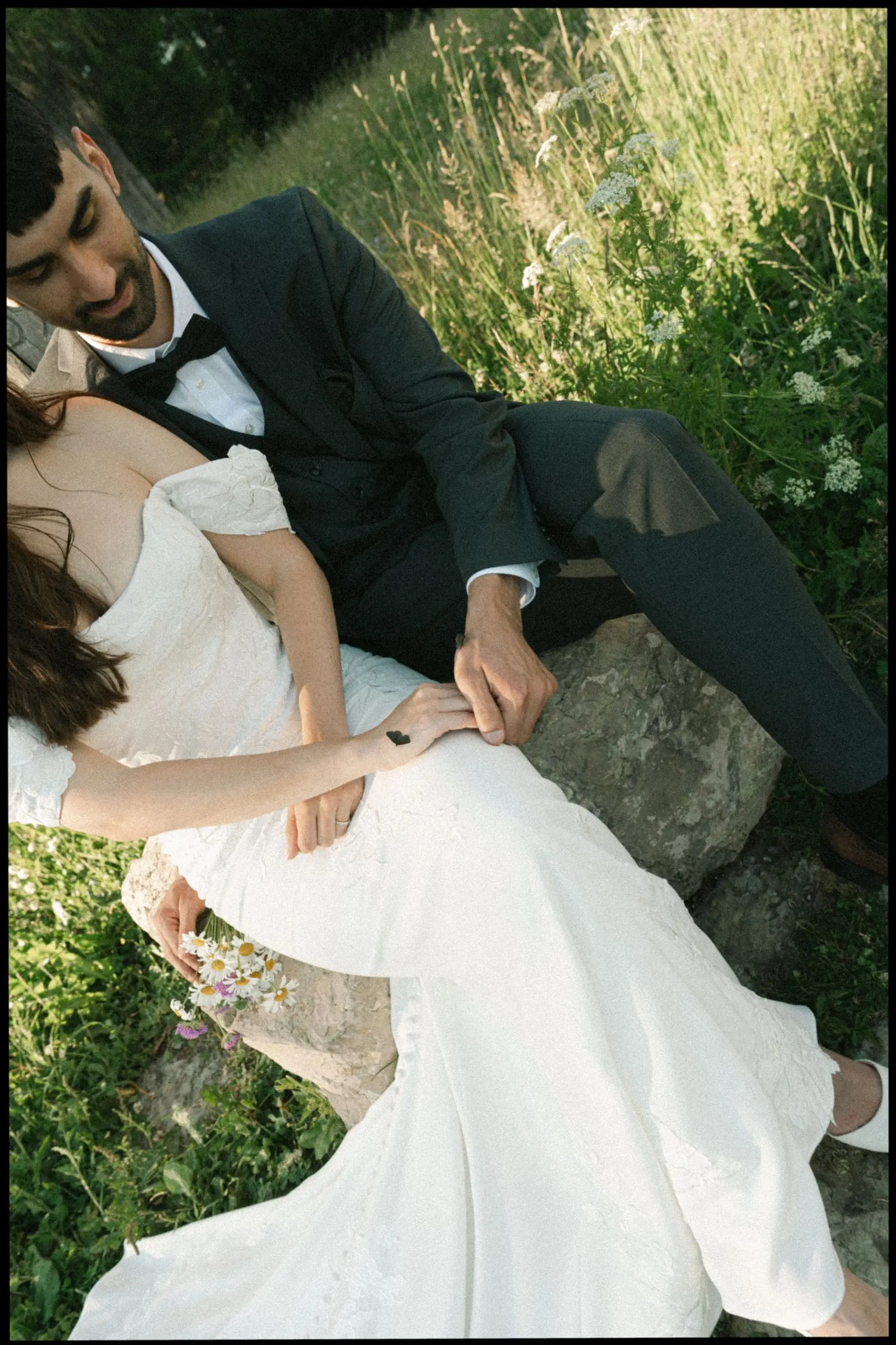 Bride resting against the groom on a rock in the tall grass, on film