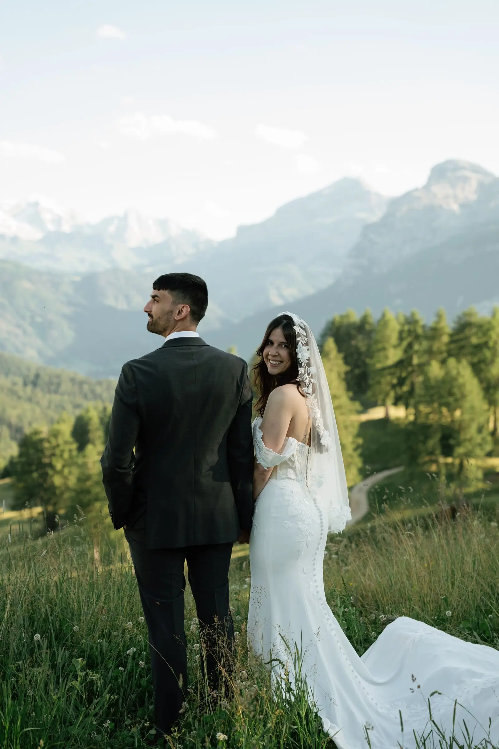Groom looks at the view as the bride smiles behind him, mountains all around