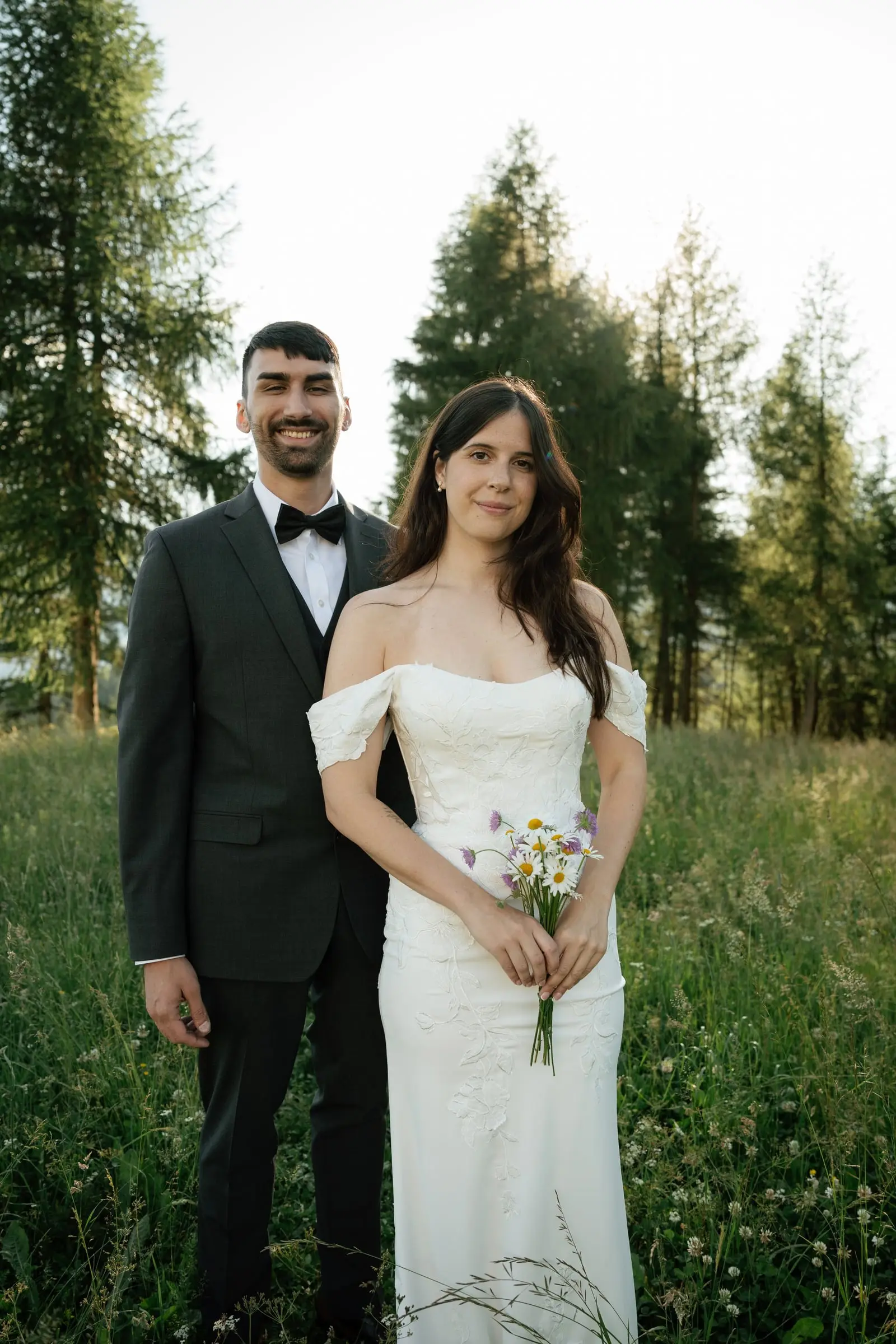 Portrait of the bride and groom in a meadow with larches, holding wildflowers