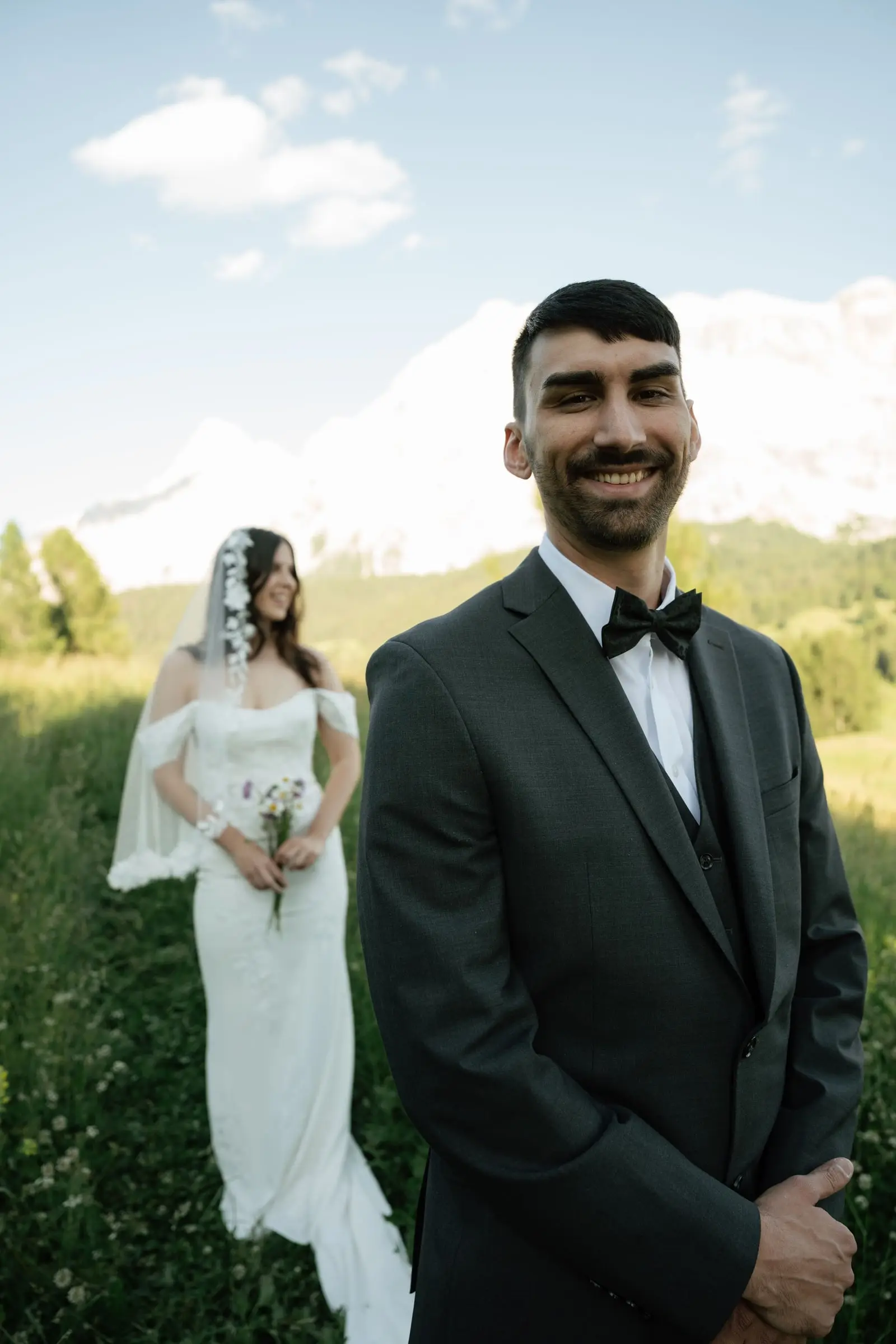 Smiling groom in the foreground, bride with veil and bouquet behind him