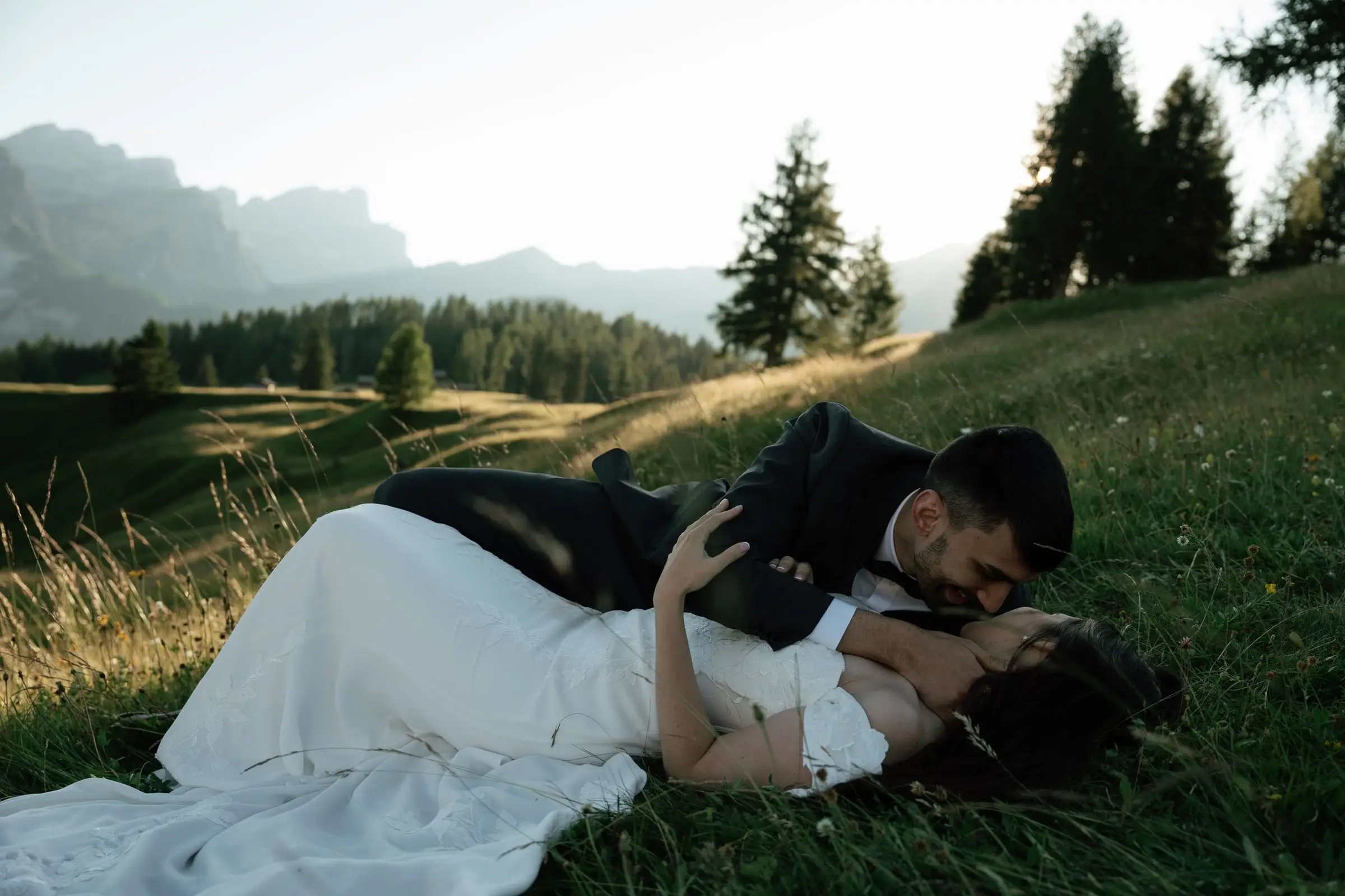 Groom kisses the bride as they lie in the meadow, peaks in the distance