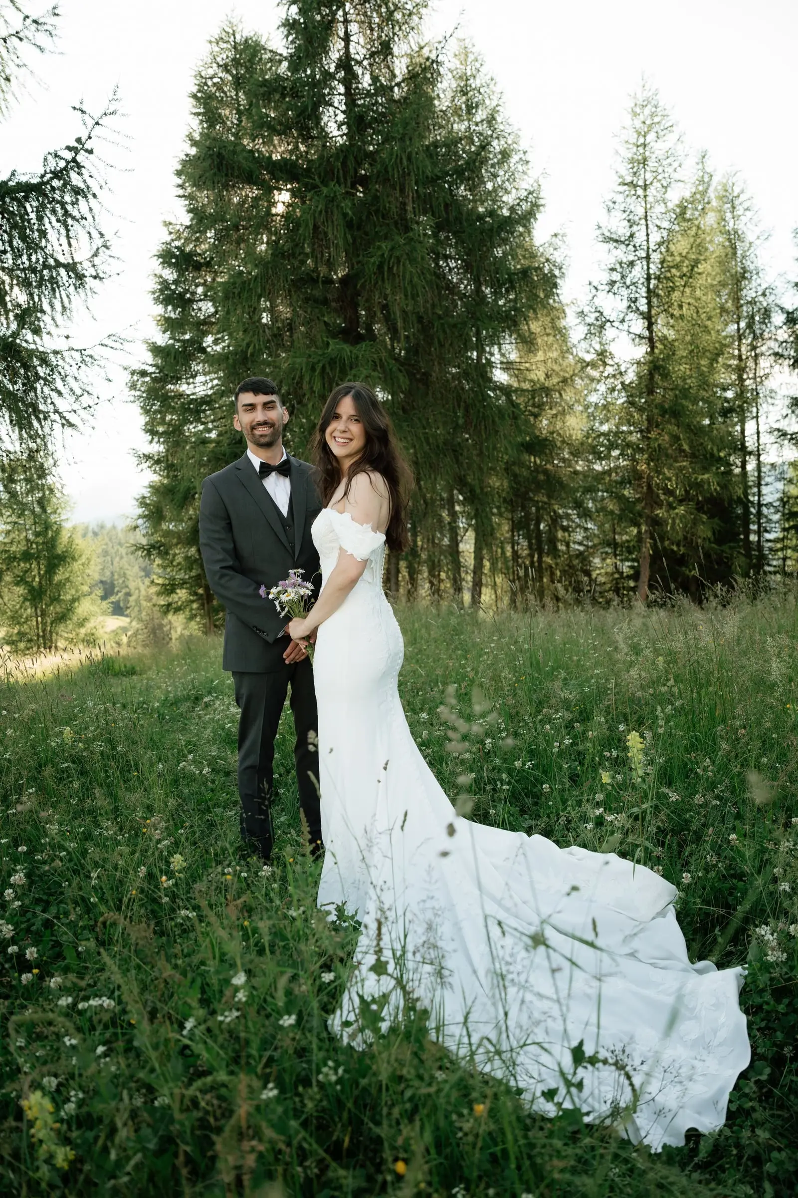 Bride with a long train standing with the groom in a meadow by the forest