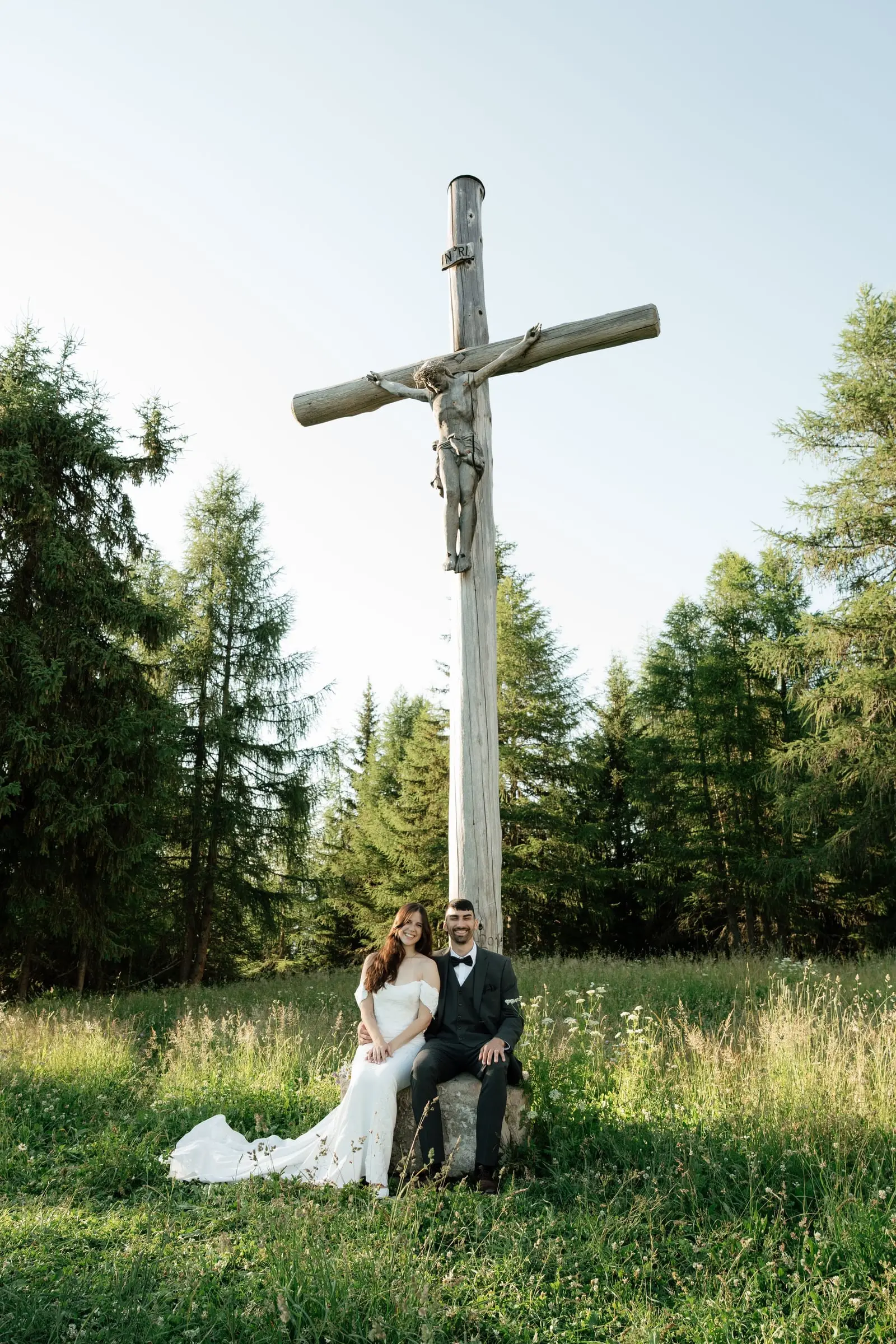 Bride and groom sitting below a tall wooden crucifix in the meadow