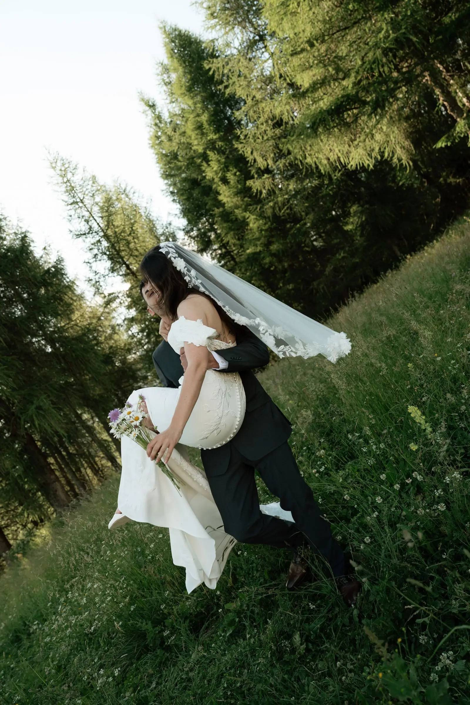 Groom carries the bride up a meadow among larches, her veil flying