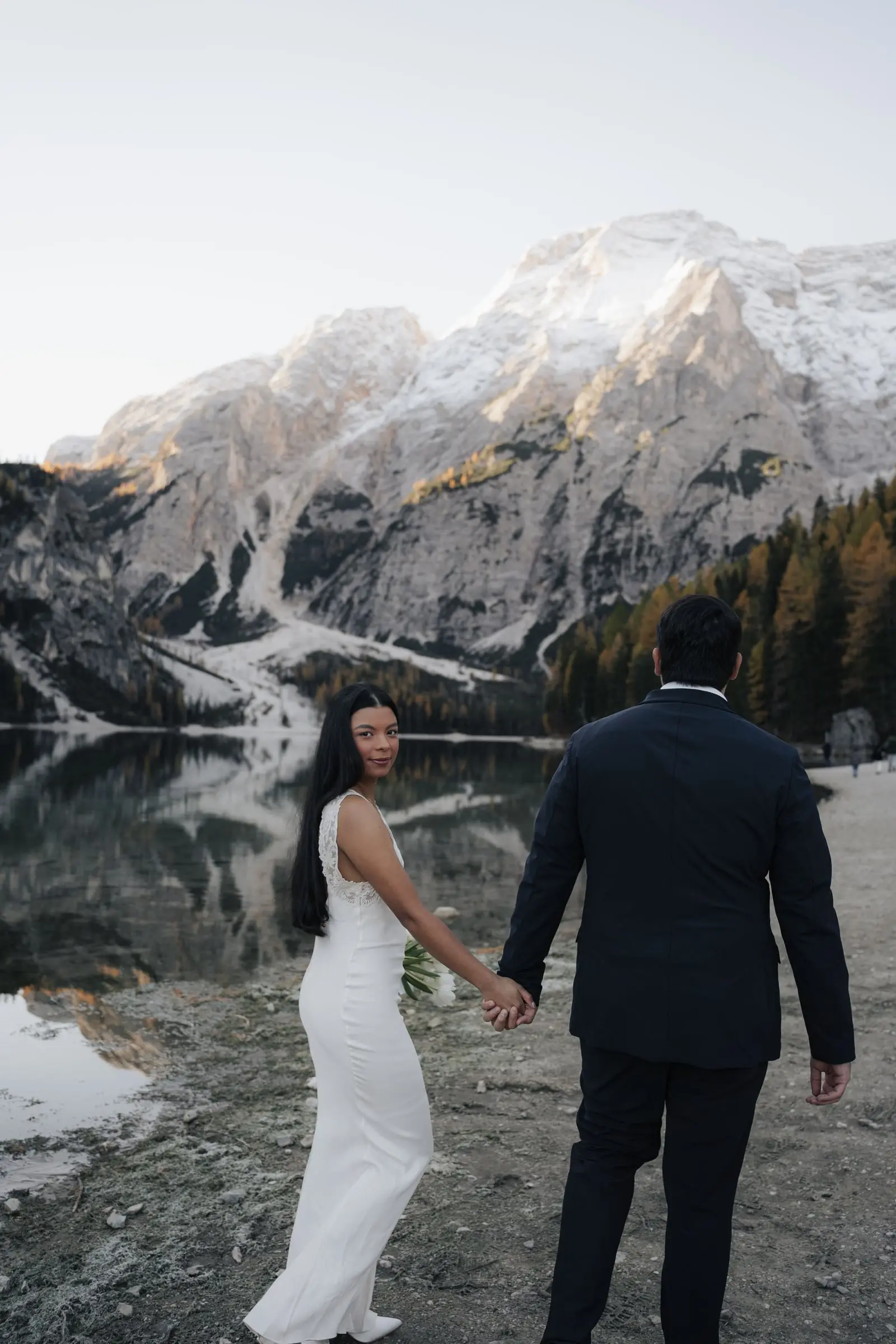 Couple walking hand in hand along the shore of Lago di Braies, snowy mountains mirrored