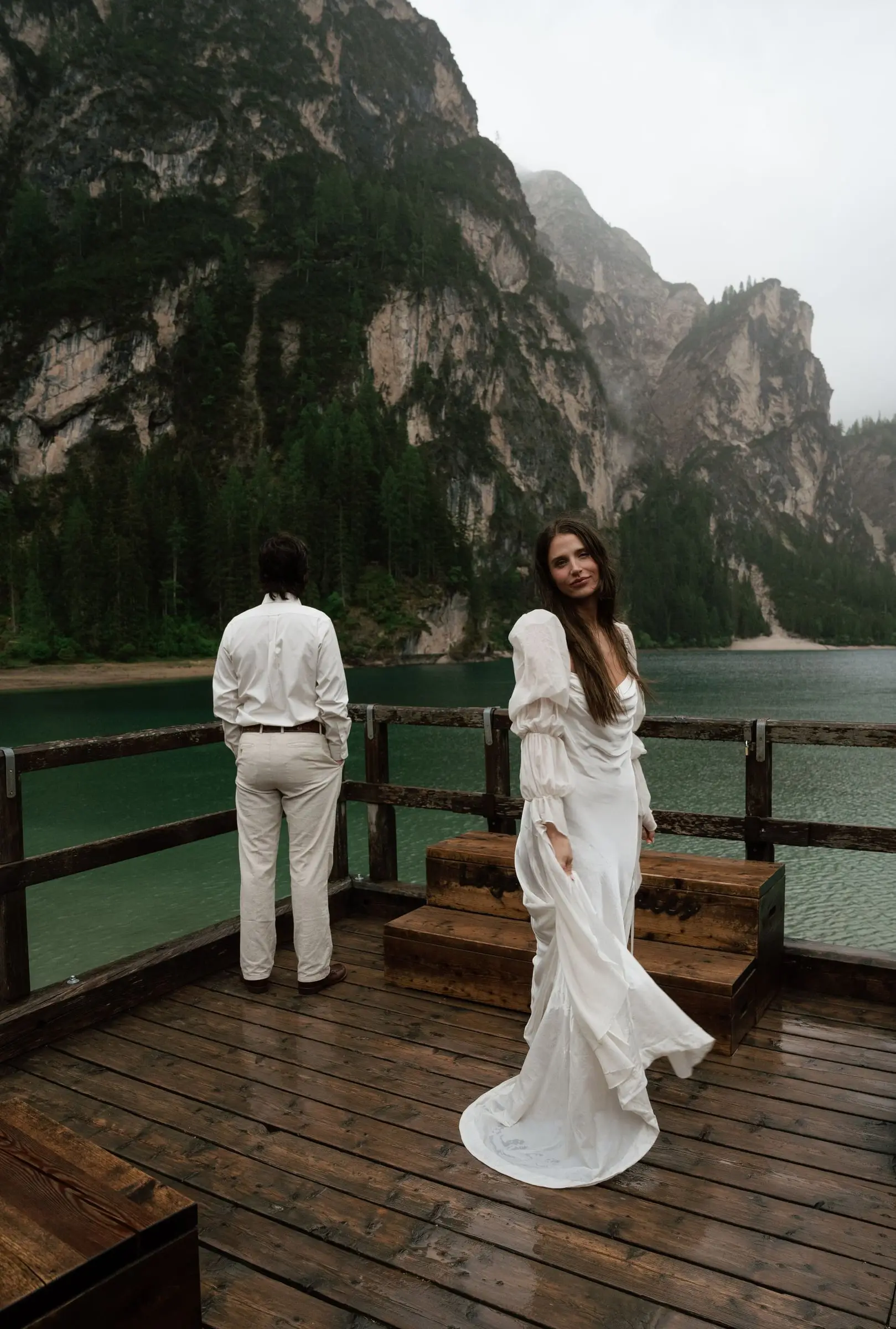 Bride in a flowing gown on the rainy boathouse terrace, the groom looking out to the lake