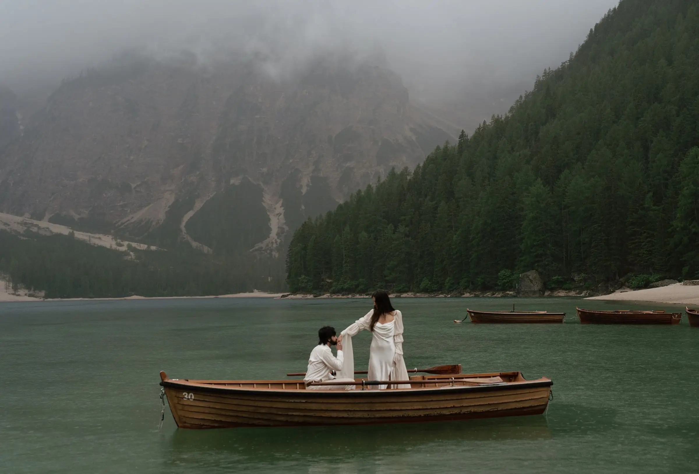 Groom kneels in a rowing boat on the misty lake, the bride standing