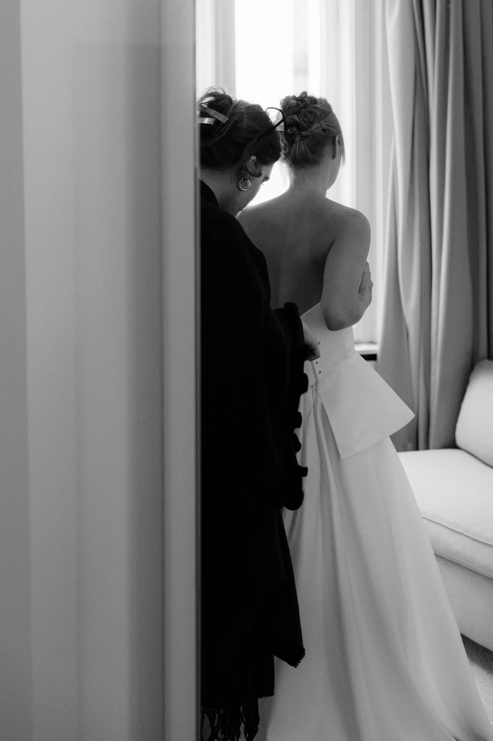 Black and white photo of the bride being helped into her gown by the window