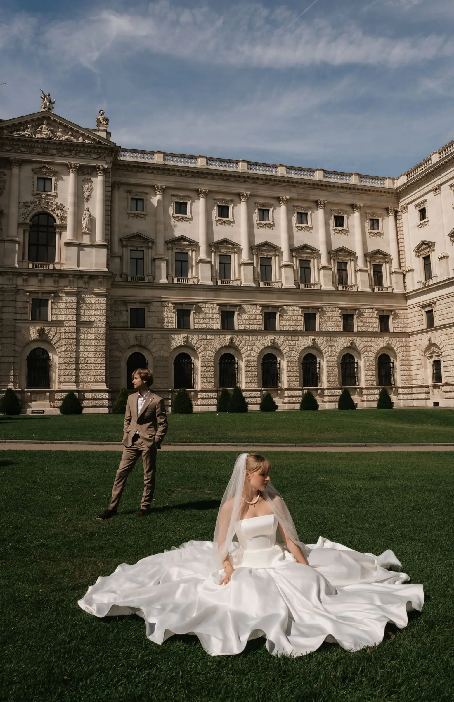 Bride sitting in her gown on the lawn in front of a Viennese palace, the groom behind