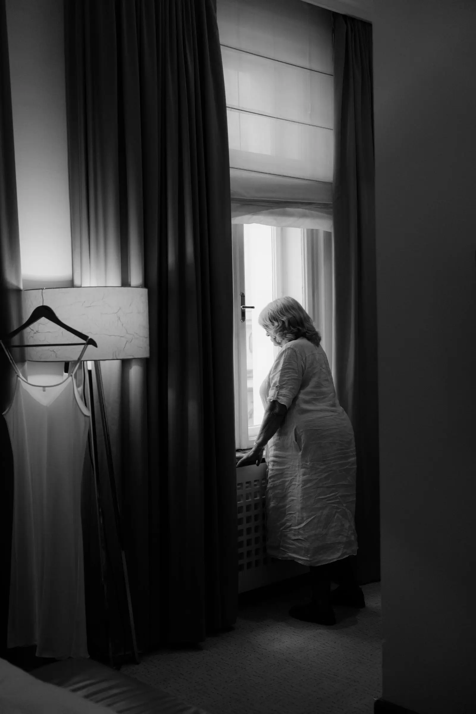 Black and white photo of the mother of the bride at the window, the dress hanging nearby
