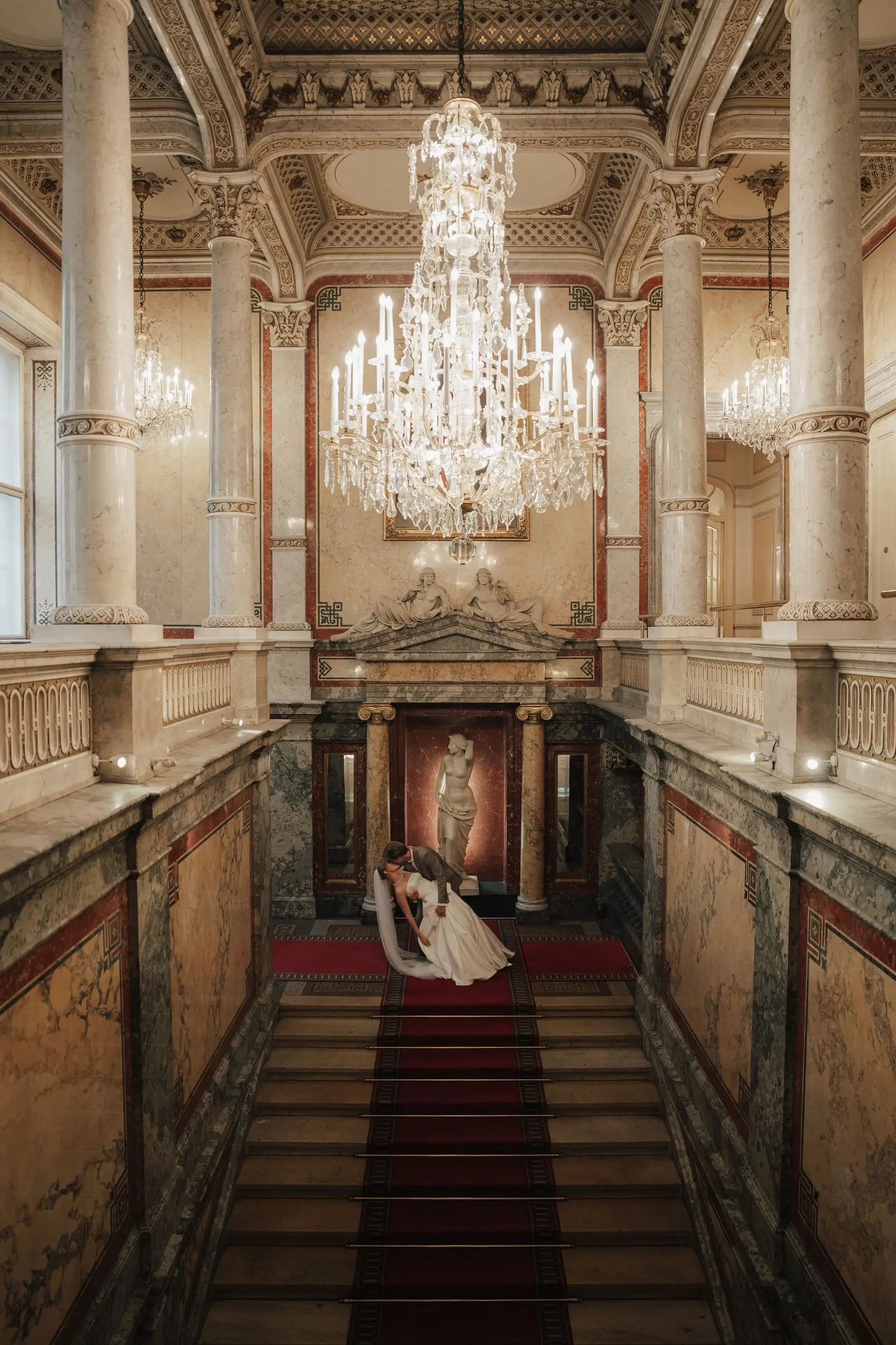 Couple kissing at the foot of the grand staircase with red carpet and chandeliers