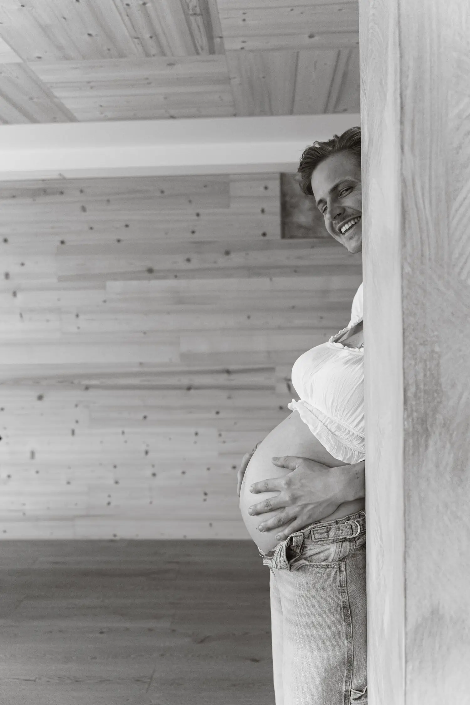 Black and white photo of the father peeking around a door at the baby bump