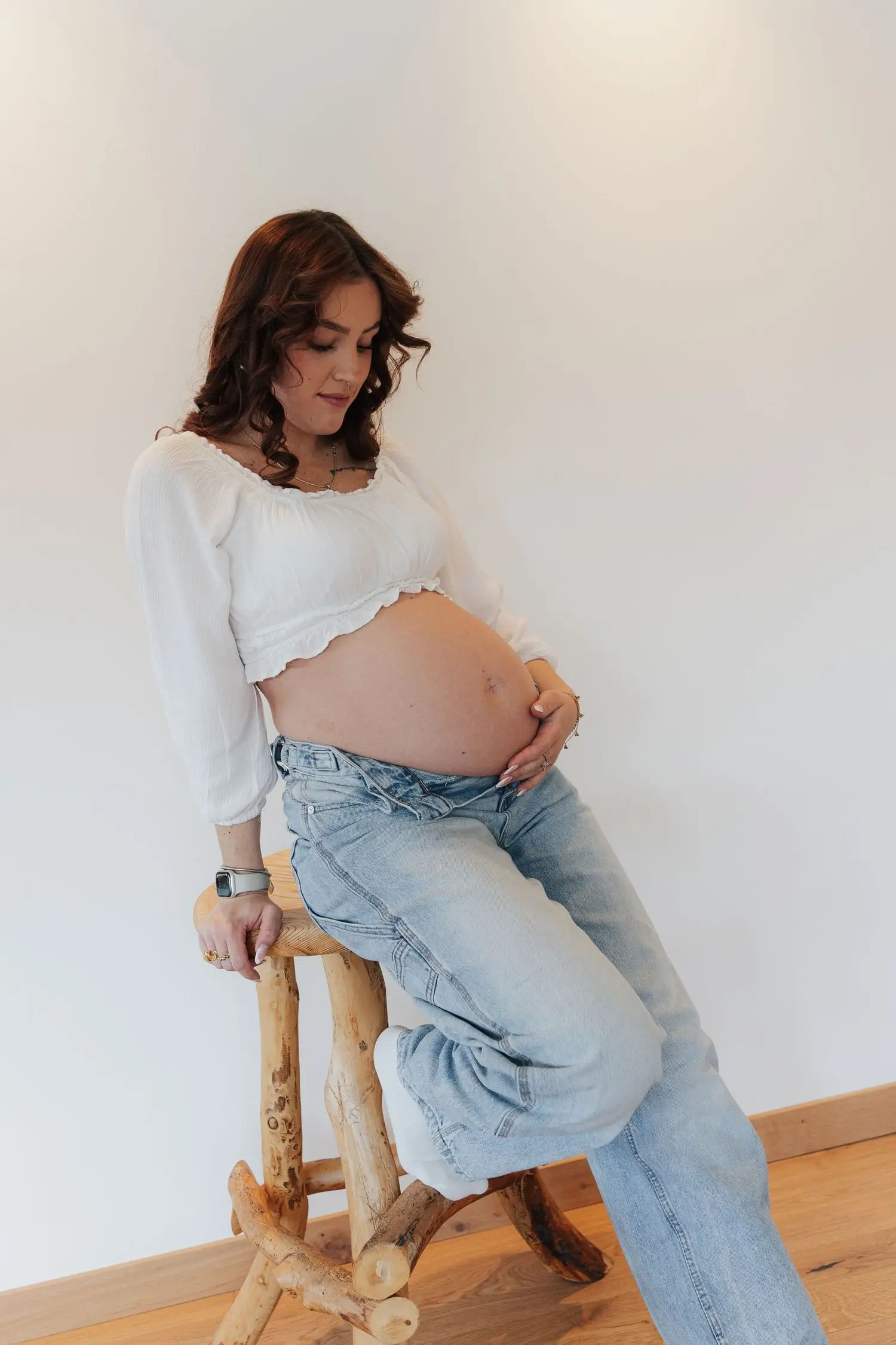 Mother-to-be in jeans and a white top sitting on a wooden stool