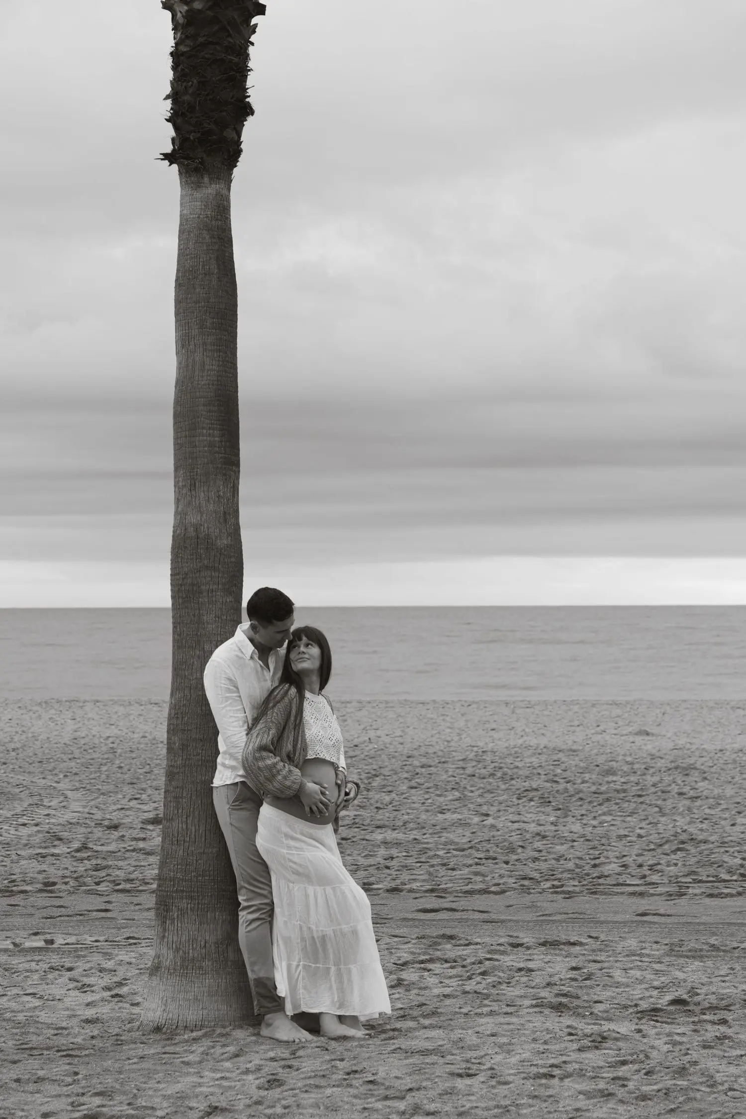 Black and white photo of the couple leaning on a palm tree at the beach