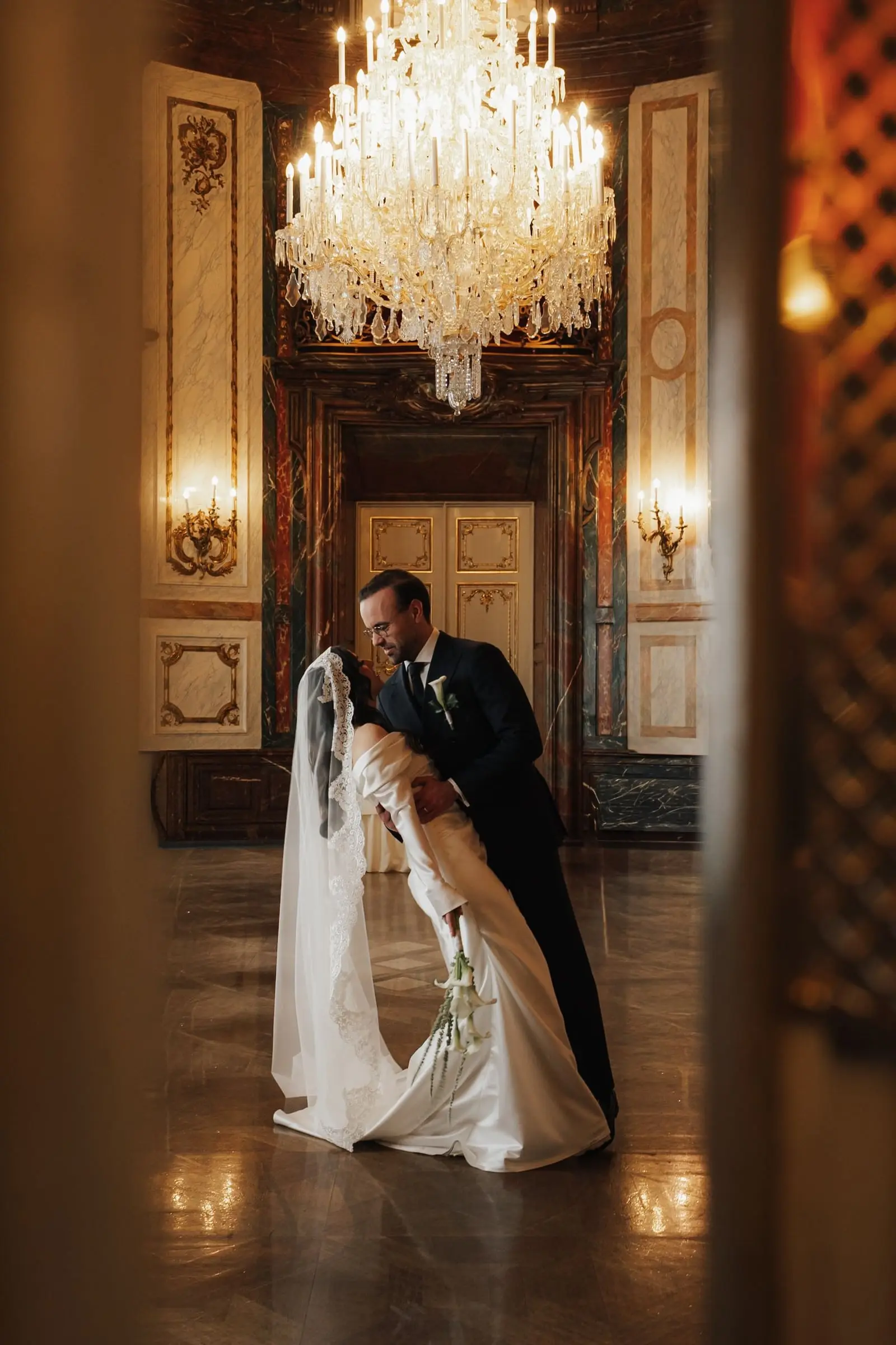Groom dips the bride under a crystal chandelier in a baroque hall of Palais Daun-Kinsky