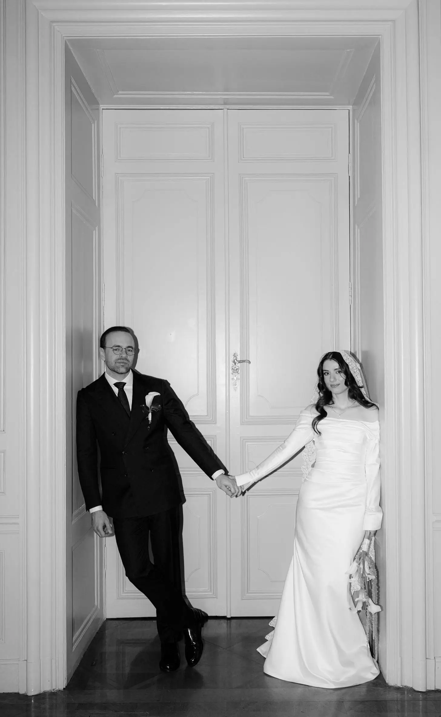 Black and white photo of the couple holding hands in a tall white doorway