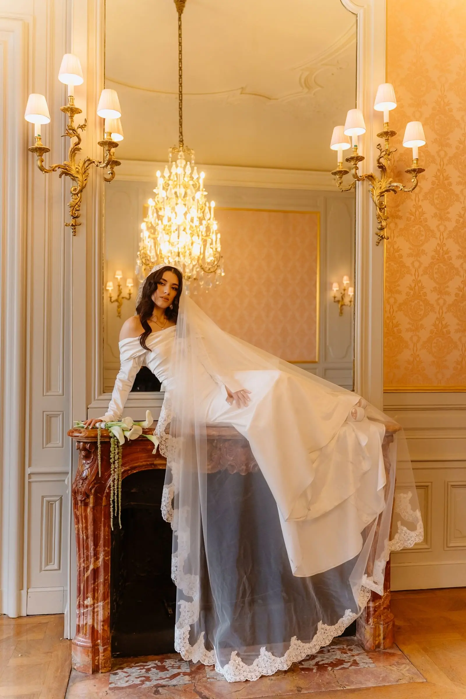 Bride standing on a marble fireplace, her long veil falling below a chandelier