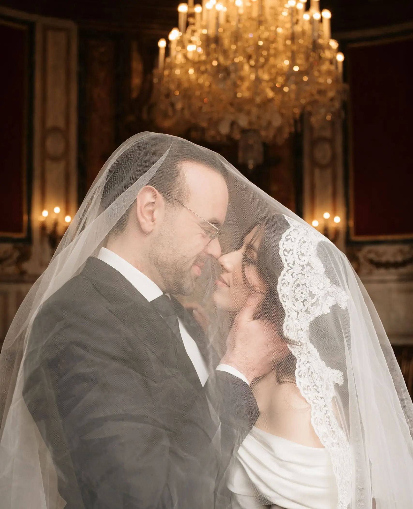 Couple under the bride's lace veil beneath a chandelier