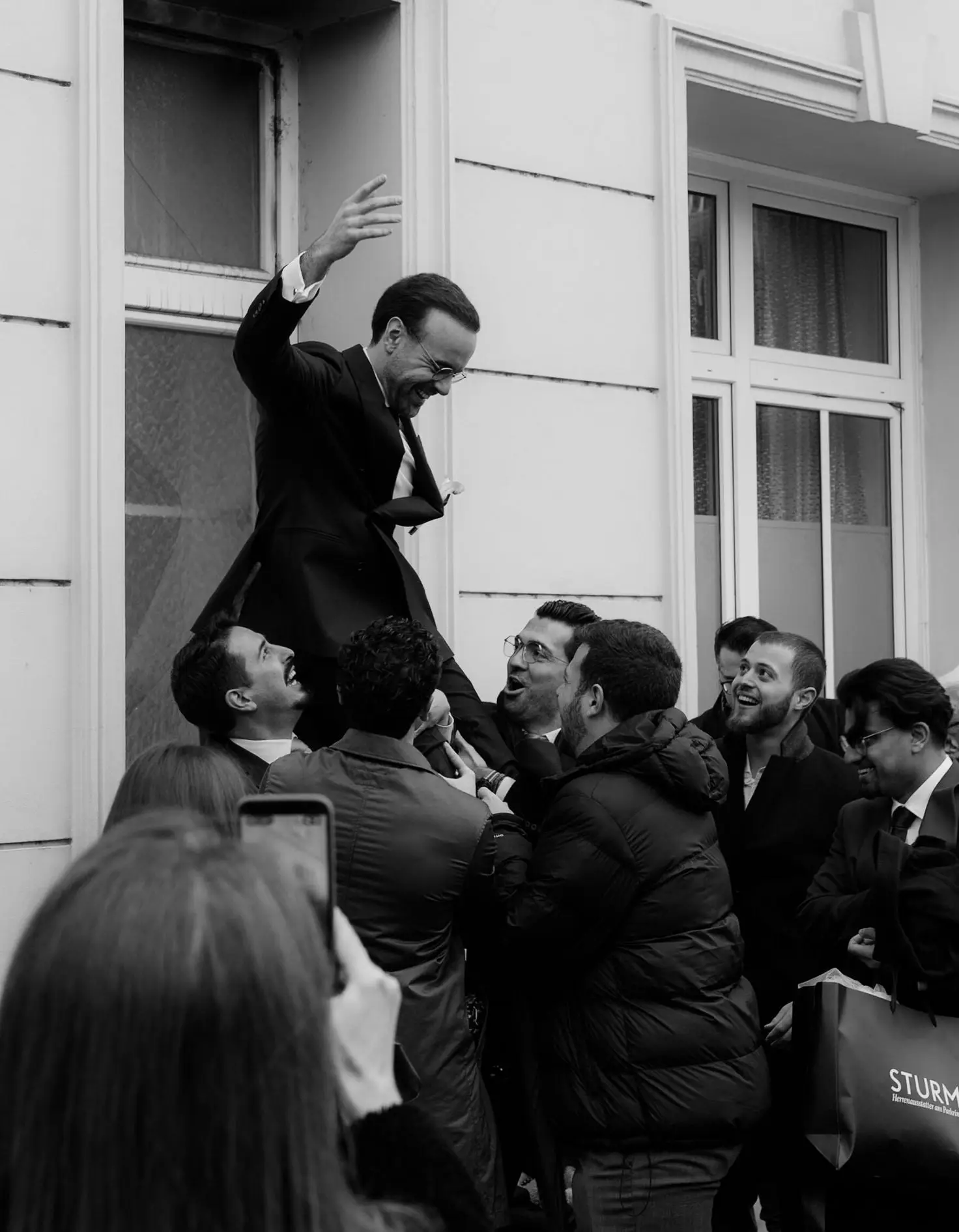 Black and white photo of guests lifting the groom in celebration