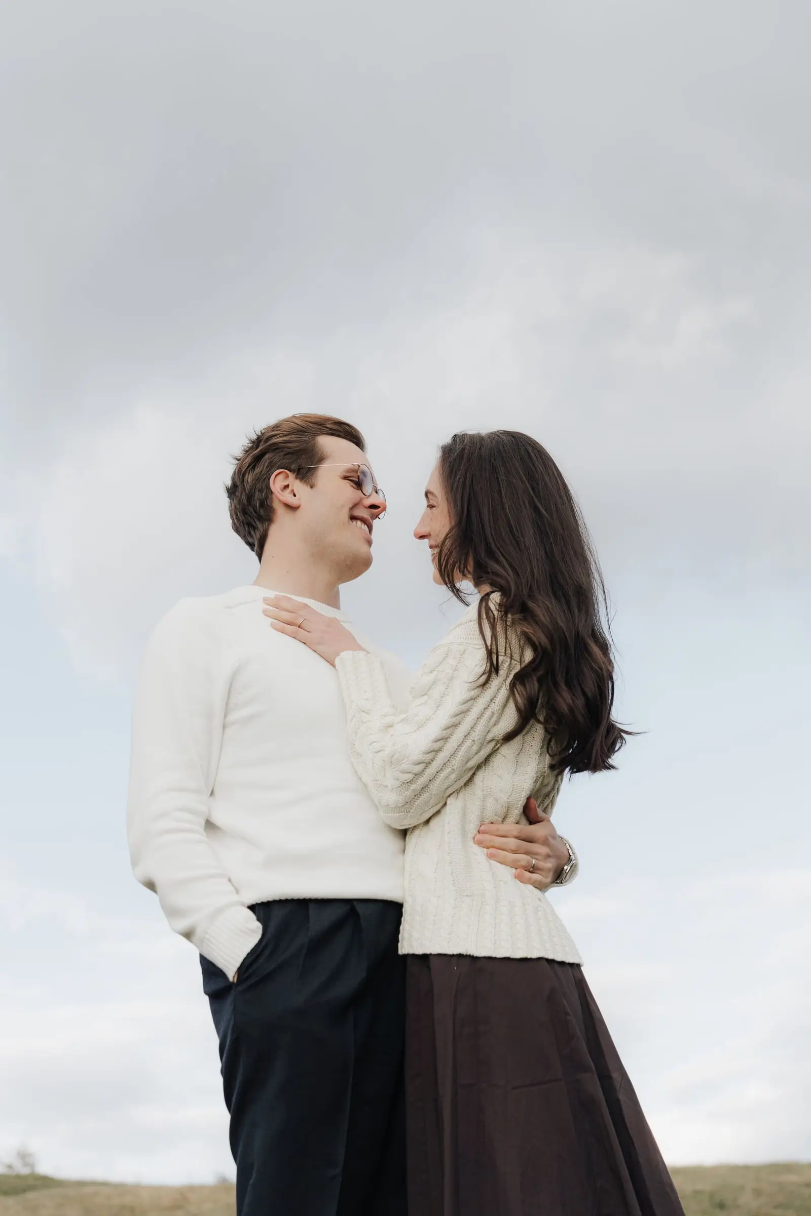Couple in cream sweaters smiling at each other against a soft sky